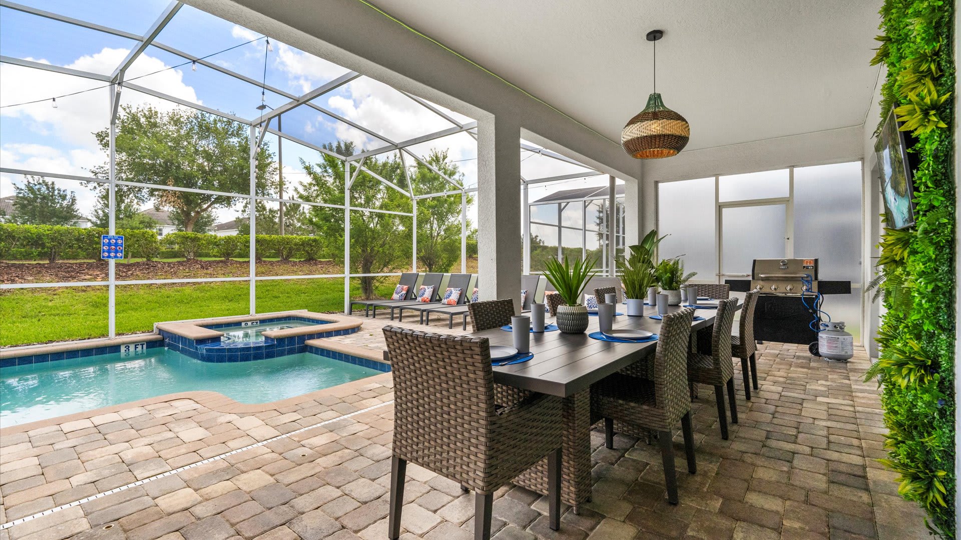 A spacious outdoor dining area under a covered lanai features a long rectangular dining table with seating for eight, set with blue place settings. A BBQ grill is visible in the background, alongside a green vertical garden wall and the private pool area.