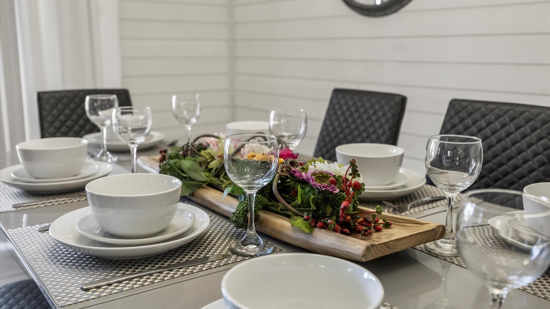 A close-up of a dining table set with elegant white dinnerware, clear wine glasses, and a vibrant floral centerpiece on a rustic wooden tray. The gray table features checkered placemats, with black quilted dining chairs visible in the background.