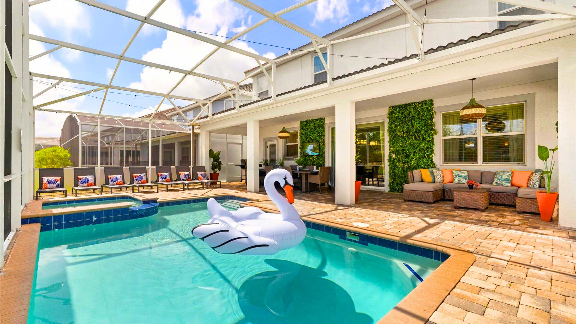 A spacious screened-in patio features a rectangular swimming pool with a large white swan float, surrounded by a brick paver deck with lounge chairs and an outdoor dining area under a covered lanai. The house exterior is visible in the background with large windows and green wall decor.