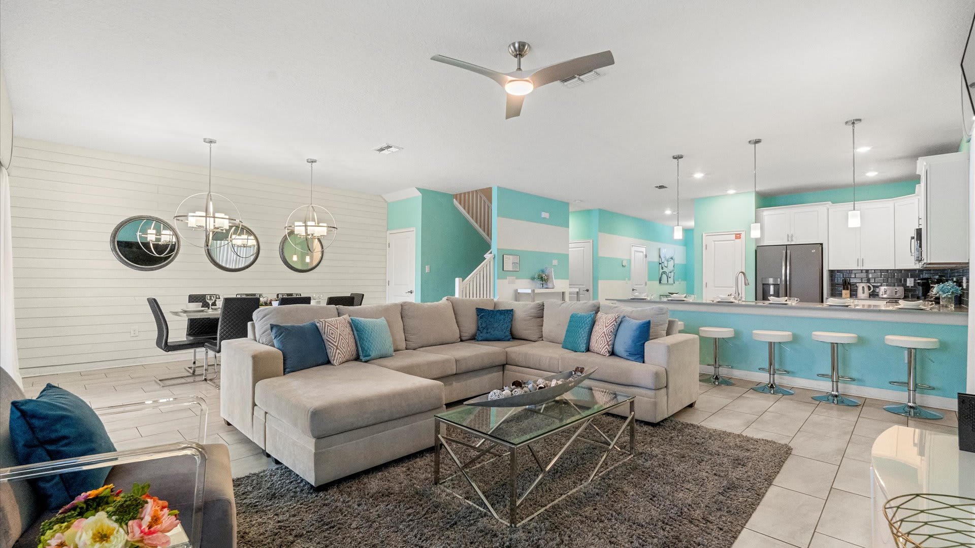 A bright, open-concept living area features a large L-shaped sectional sofa with blue accent pillows, a glass coffee table, and a gray area rug, with a dining area and kitchen bar visible in the background.