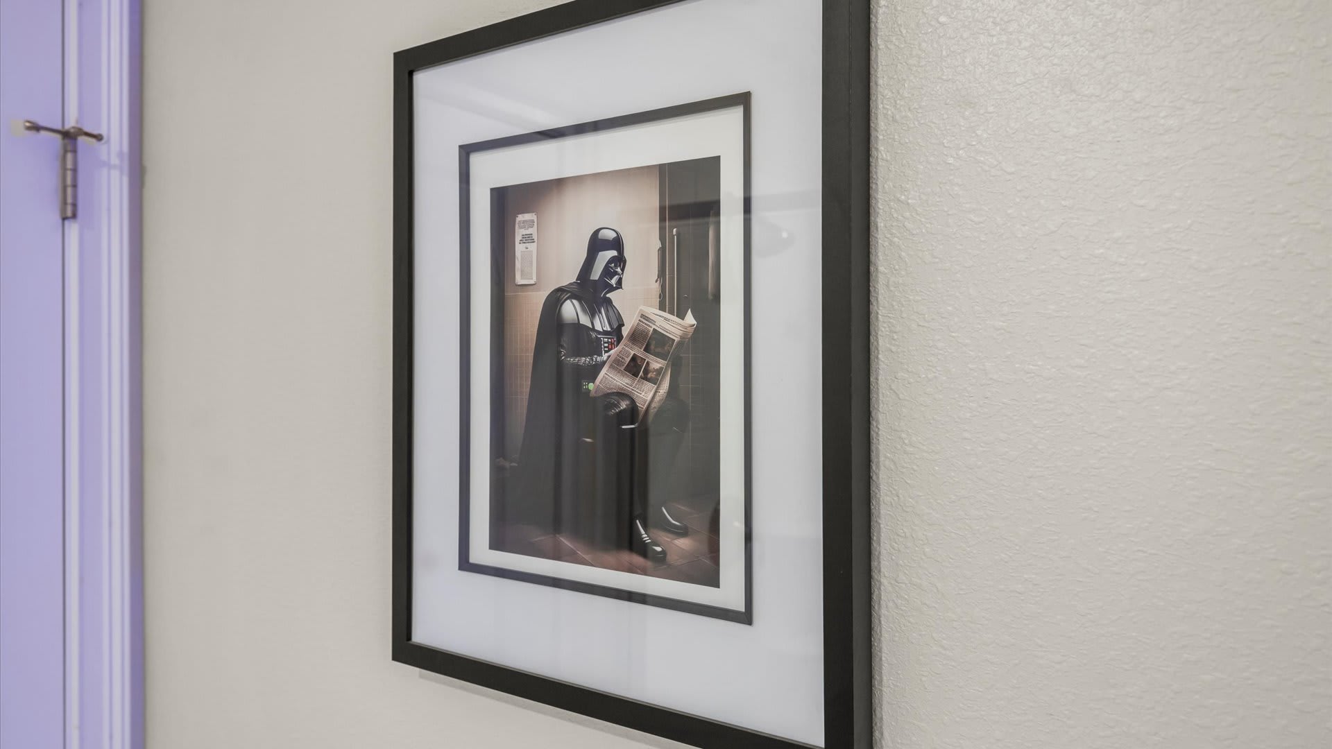 A humorous framed artwork depicting Darth Vader sitting on a toilet and reading a newspaper, set against a light-colored wall.
