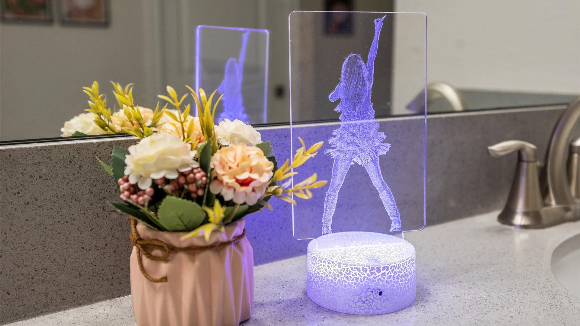 A close-up of a bathroom vanity countertop featuring a small vase of artificial yellow and pink flowers next to a clear acrylic lamp illuminating a figure with purple light.