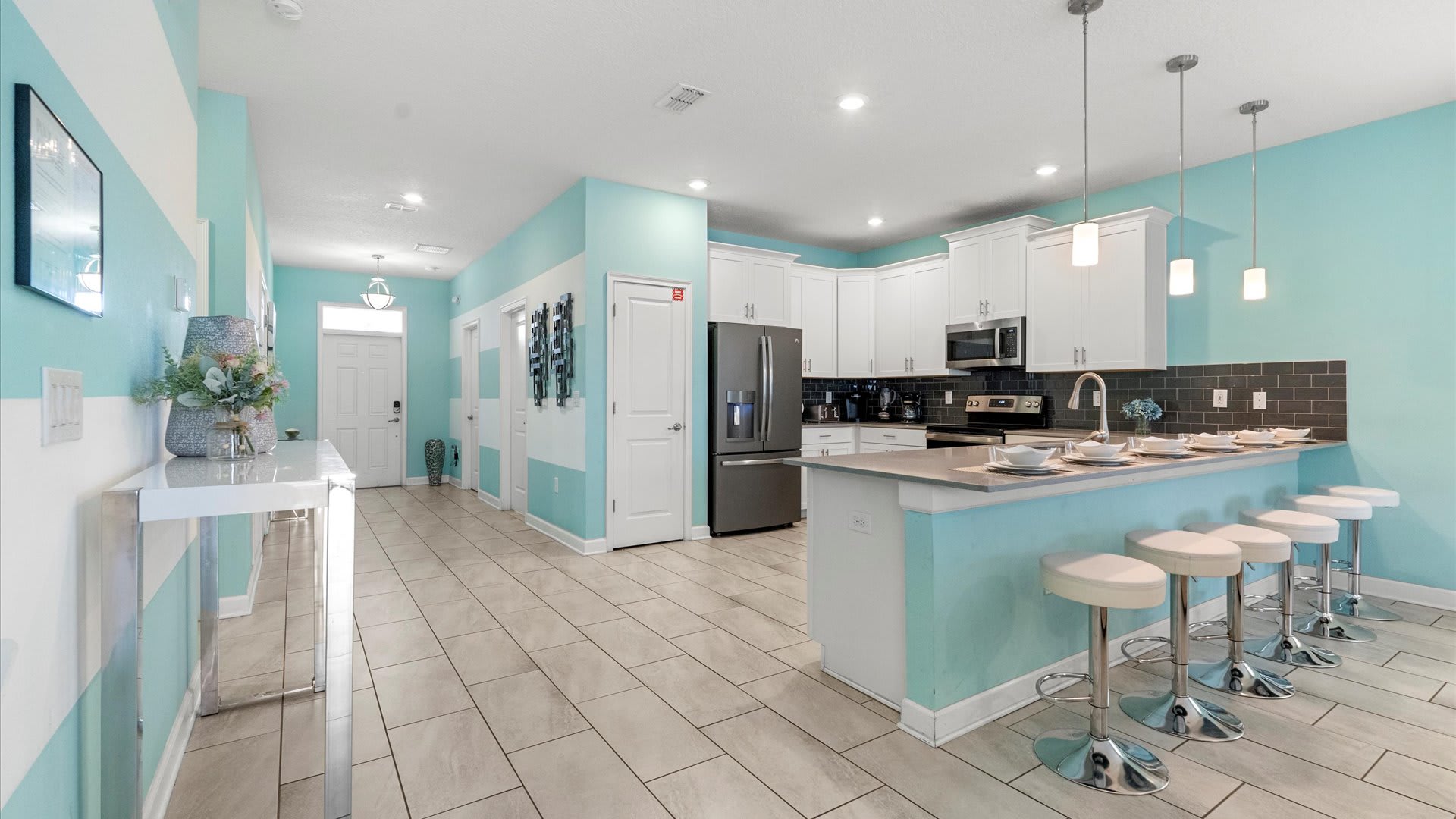 A spacious open-concept kitchen boasts white shaker cabinets, a dark subway tile backsplash, and light gray countertops. A large island with bar seating and pendant lights separates the kitchen from the living area, featuring vibrant aqua walls.