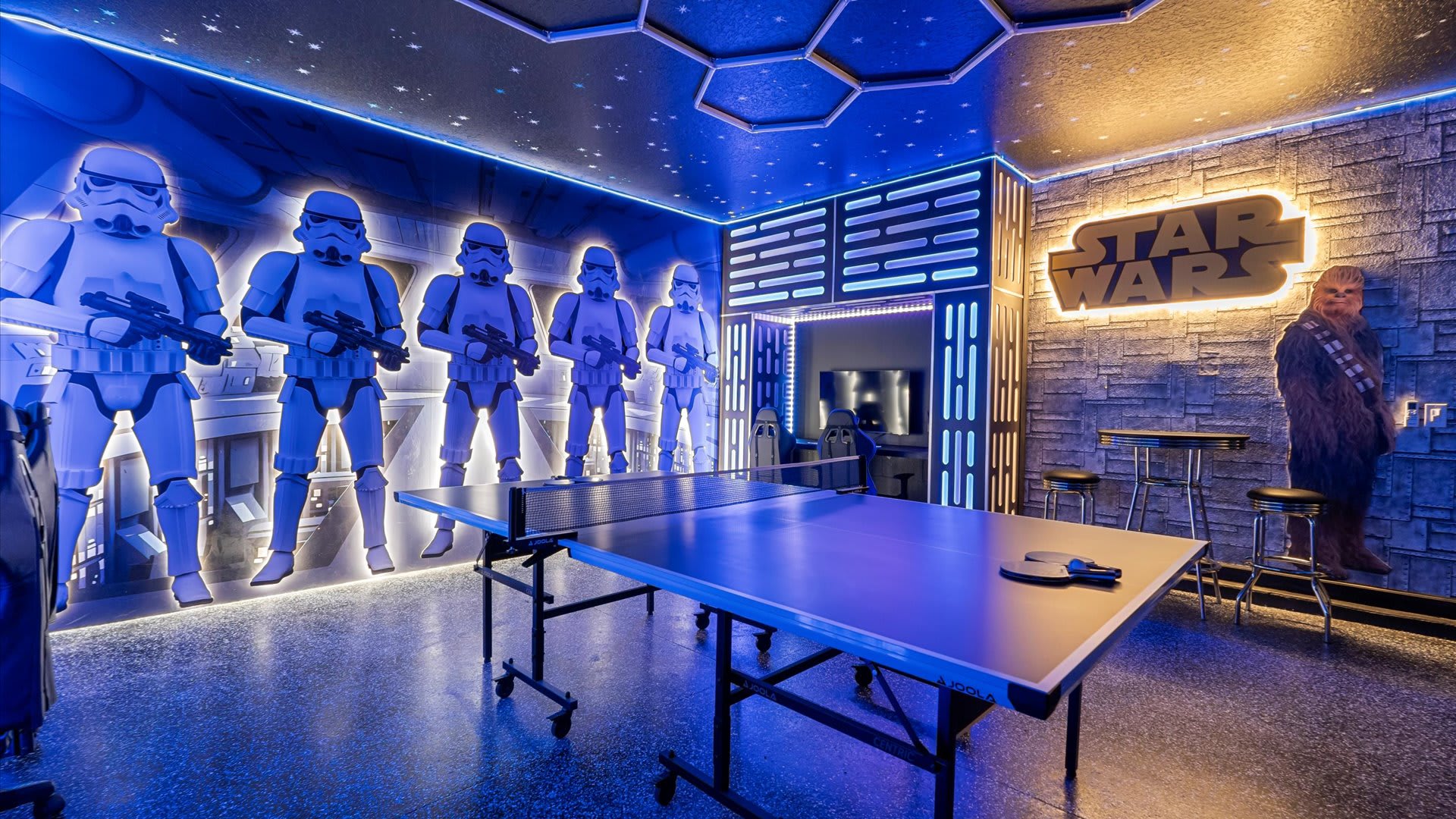 A vibrant Star Wars-themed game room features a ping-pong table in the center, surrounded by life-sized Stormtrooper figures, a Chewbacca figure, and glowing blue and white LED lighting accents on the walls and ceiling.