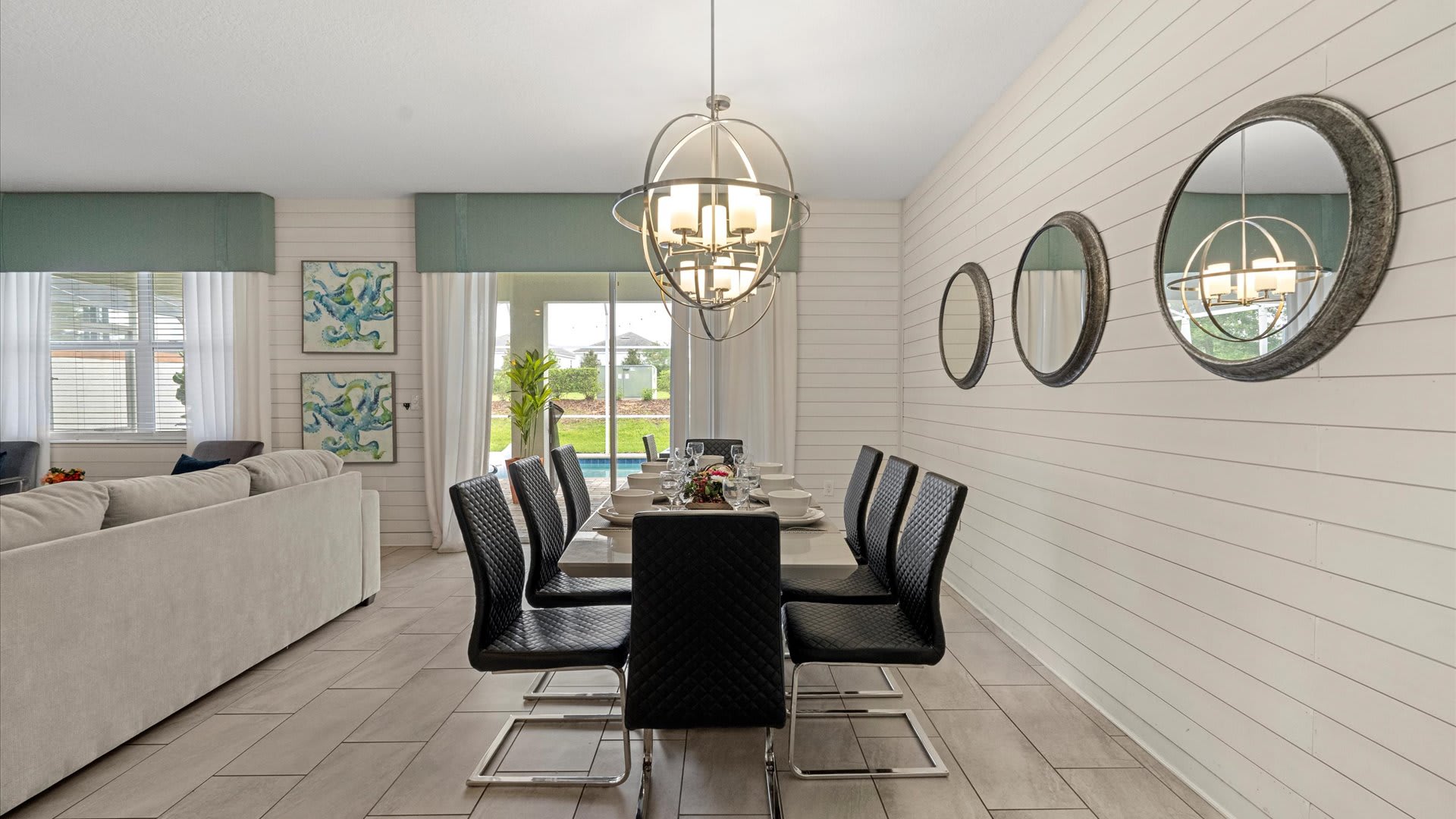 An open-concept dining room features a long gray table with eight black quilted chairs, positioned adjacent to a large light gray sectional sofa. Two spherical chandeliers hang above the table, and white shiplap walls are decorated with round mirrors.