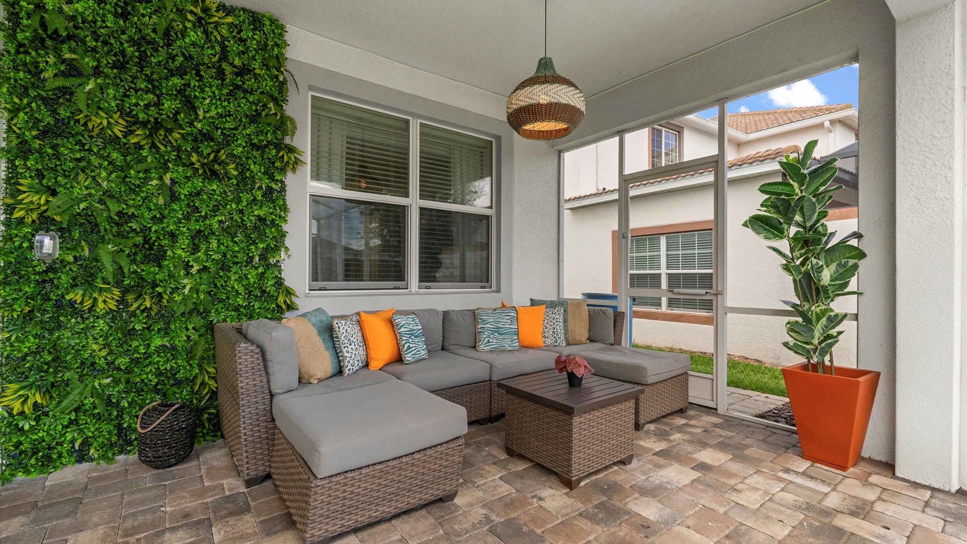A cozy outdoor lounge area on a covered patio features a large L-shaped wicker sofa with colorful throw pillows and a matching coffee table. A vibrant green vertical garden wall and a tall potted plant add natural elements to the space.