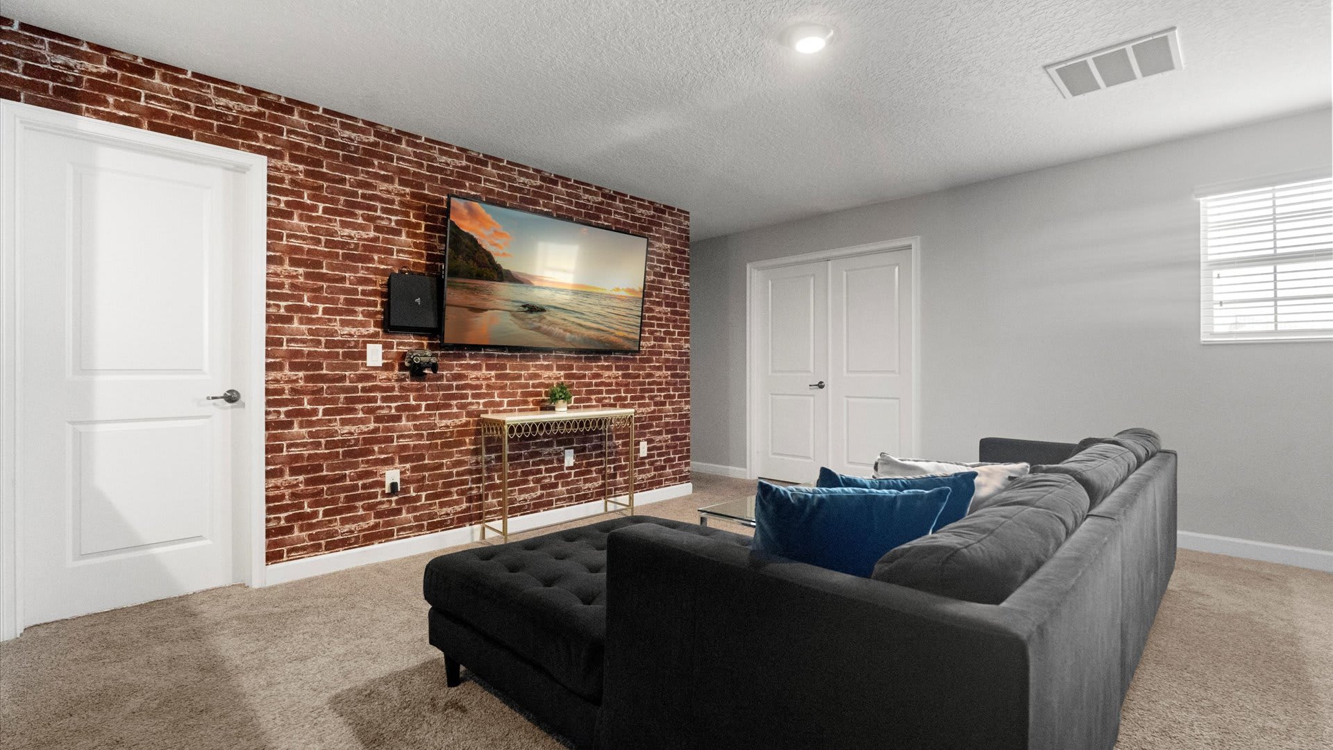 This media room features a dark gray sectional sofa with blue throw pillows, facing a wall-mounted TV on a red brick accent wall, with a small window and white closet doors visible on the light gray wall.