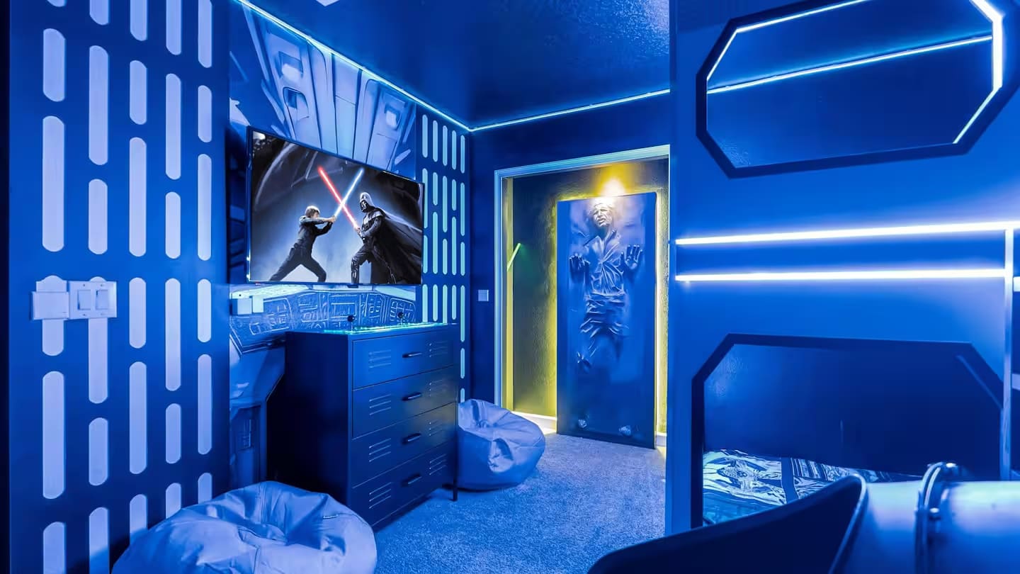A unique Star Wars-themed bedroom features a wall-mounted TV displaying a lightsaber duel, two gray bean bag chairs, and a dark blue dresser, all under blue LED strip lighting and a dark ceiling with star-like lights.