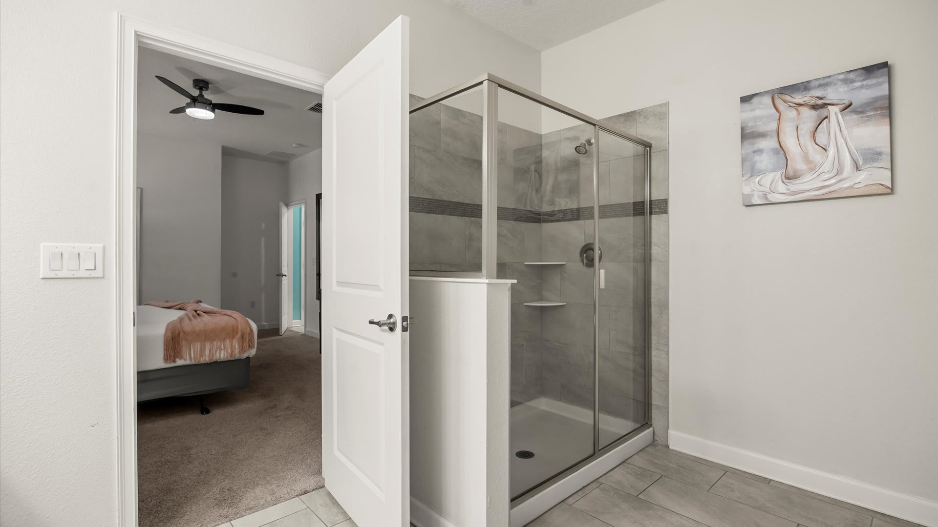 A modern bathroom with a glass-enclosed walk-in shower featuring gray tiled walls and a decorative horizontal stripe. The view through the open door reveals a glimpse of a bedroom with a bed and a peach throw.