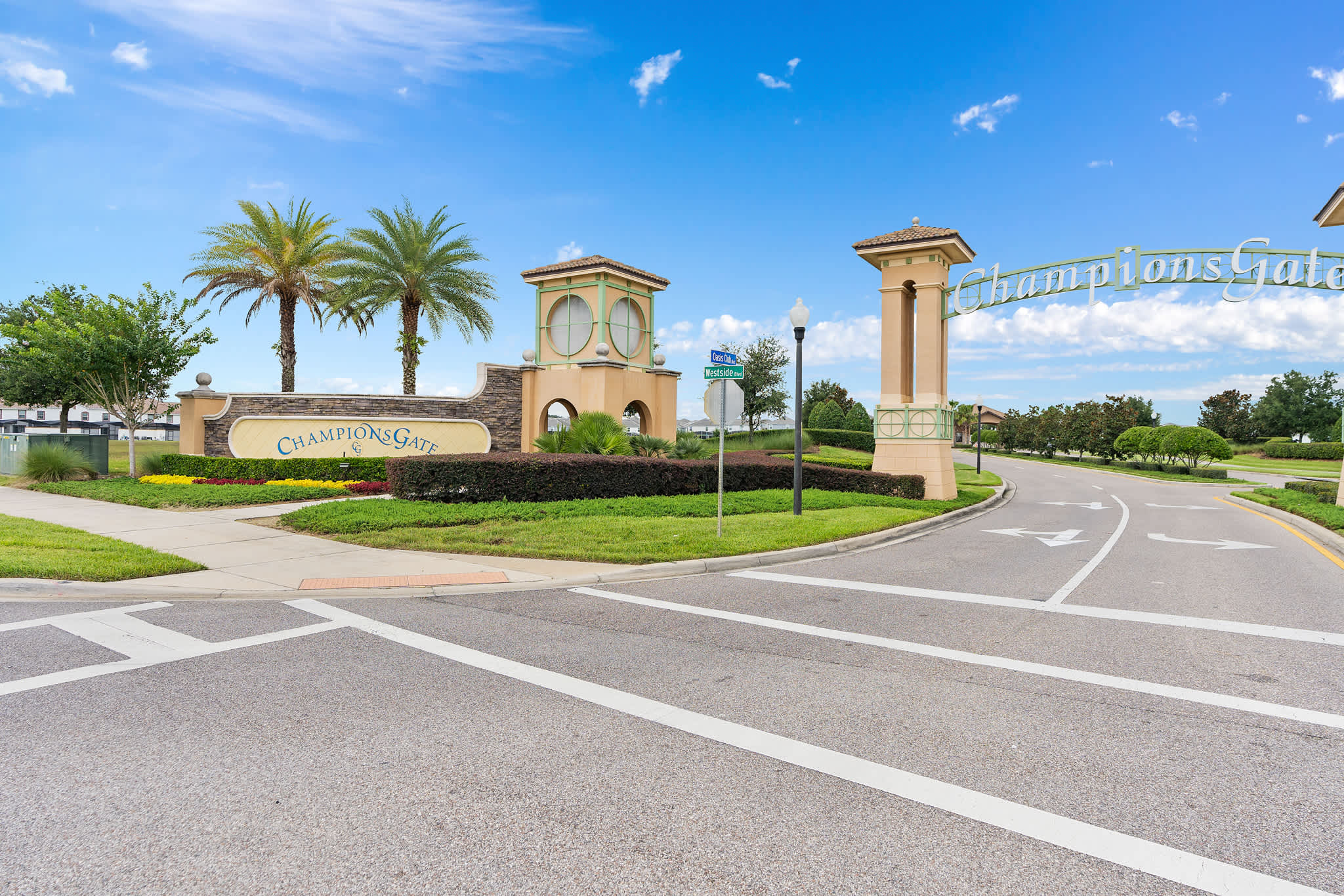 A grand entrance to the "ChampionsGate" community features large stone pillars with ornate detailing, lush green landscaping, and tall palm trees under a bright blue sky. A multi-lane road leads into the community, indicating a significant development.