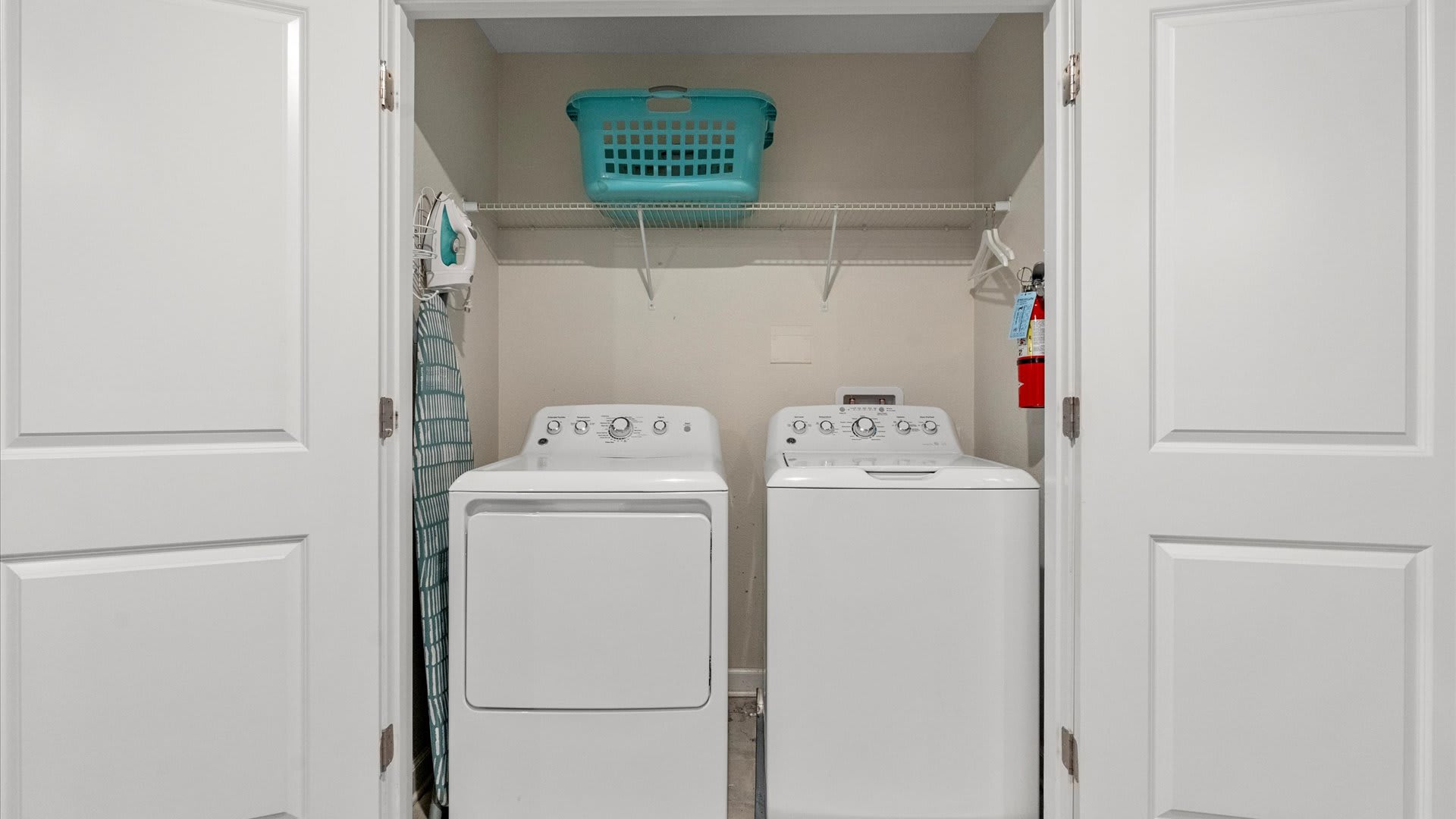 A compact laundry closet featuring a white top-load washer and front-load dryer, an ironing board with an iron, and a blue laundry basket on a wire shelf above.