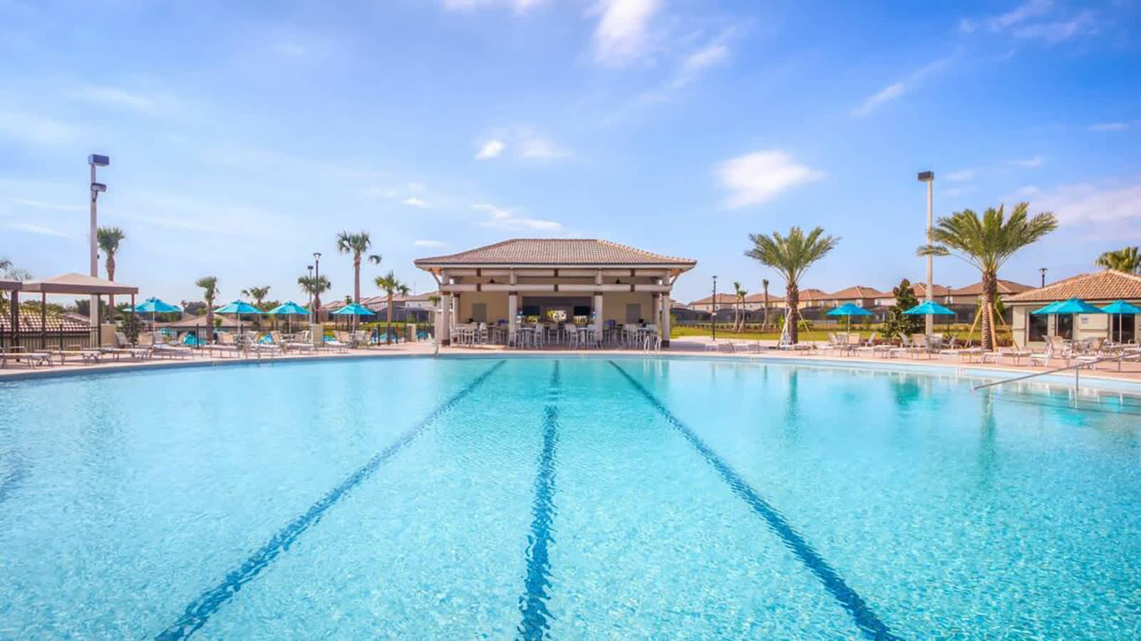 A spacious, sparkling blue communal swimming pool with lane markers stretches towards a covered outdoor bar area in the background, surrounded by lounge chairs and palm trees under a clear sky. The pool appears ideal for both relaxation and swimming laps.