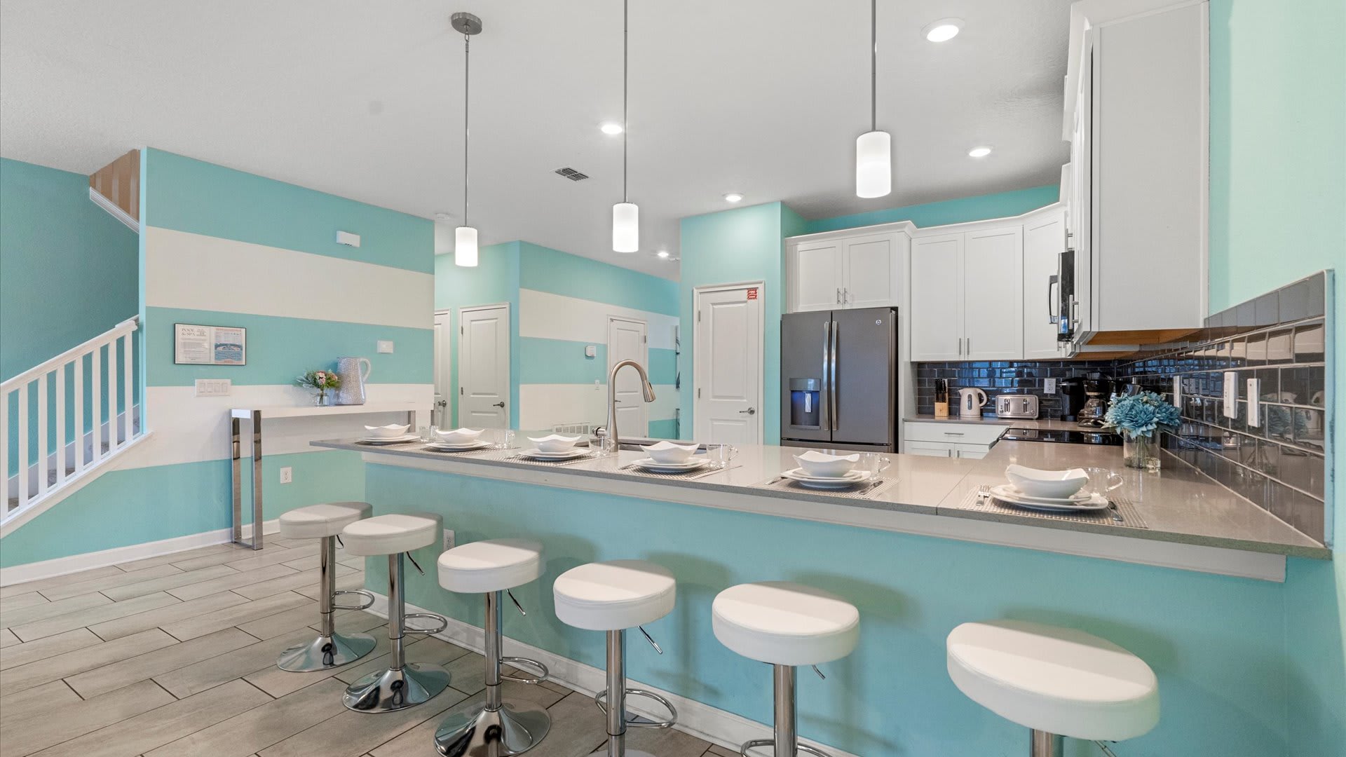 A modern kitchen features a long island with five white bar stools, light gray quartz countertops, white shaker cabinets, and a dark gray subway tile backsplash, all set against a vibrant turquoise and white striped accent wall.