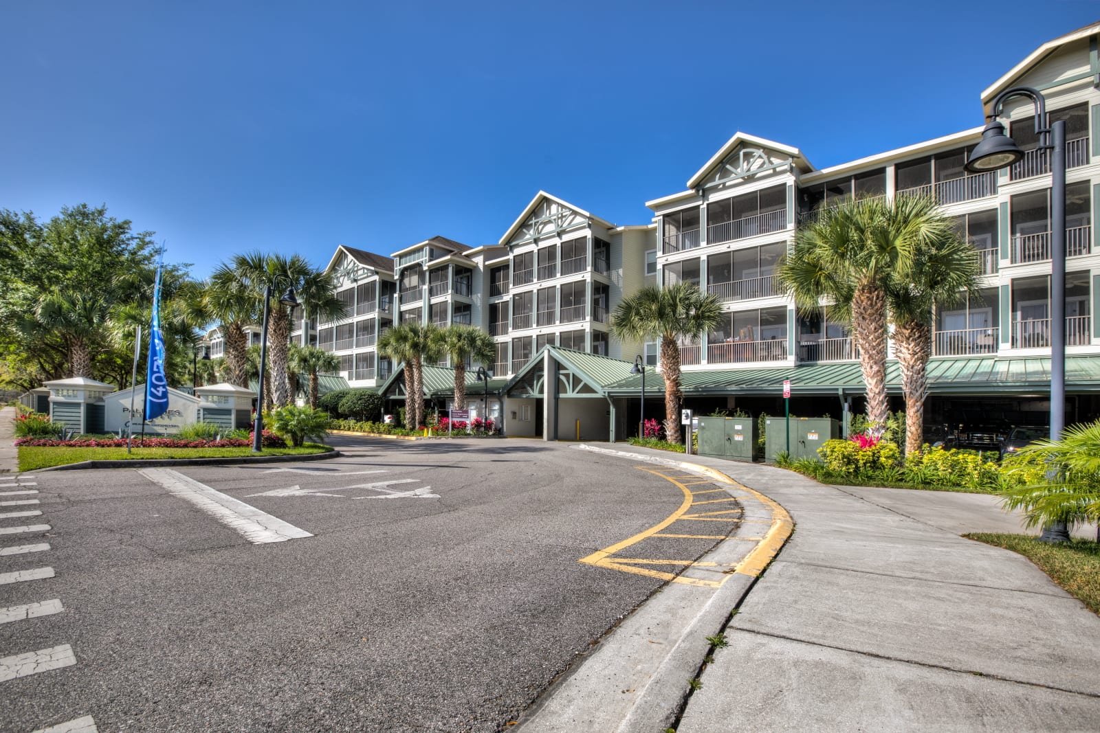 The grand front entrance of a multi-story condominium building, characterized by green roofs, screened balconies, and lush palm trees under a bright blue sky.