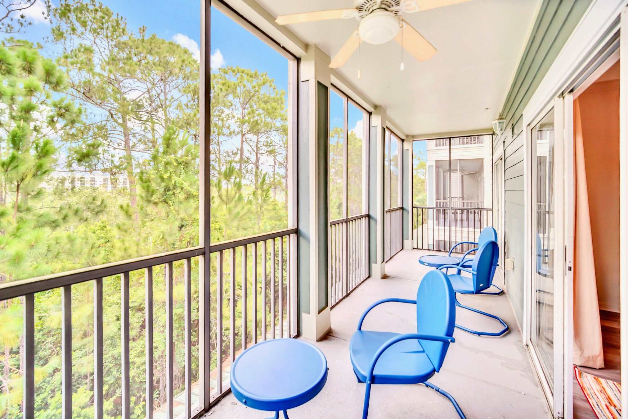 A wide-angle view of a screened-in balcony furnished with blue metal chairs and a small table, overlooking a dense canopy of pine trees under a bright blue sky.