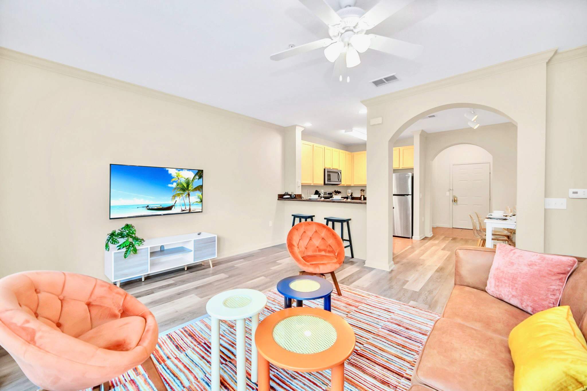 An open-concept living area with light wood-look flooring, a brown sofa, and two orange armchairs on a striped rug, facing a wall-mounted TV, with a breakfast bar and kitchen visible in the background.