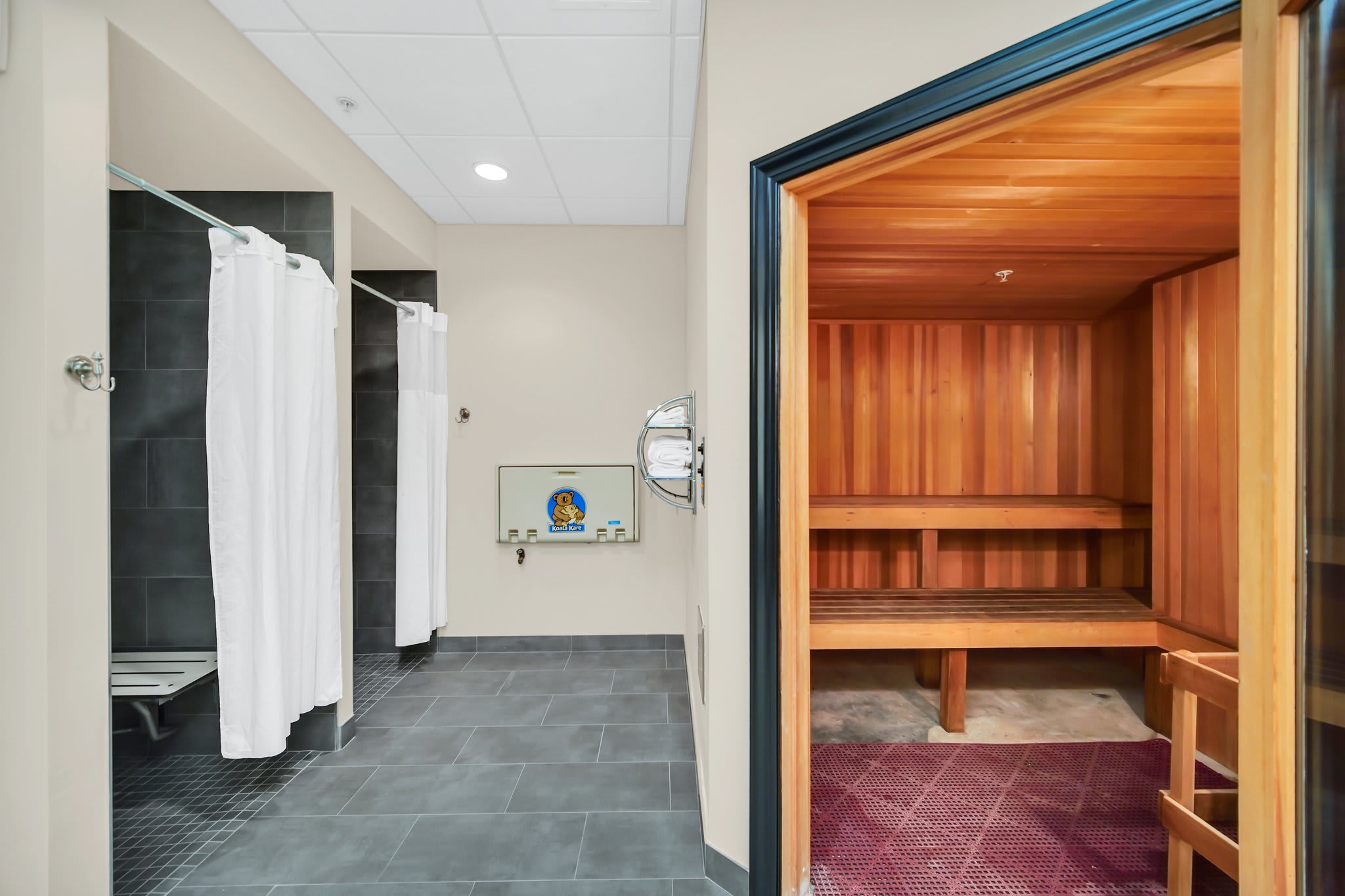 A clean changing area with multiple shower stalls featuring white curtains, adjacent to a traditional wooden sauna room with benches.