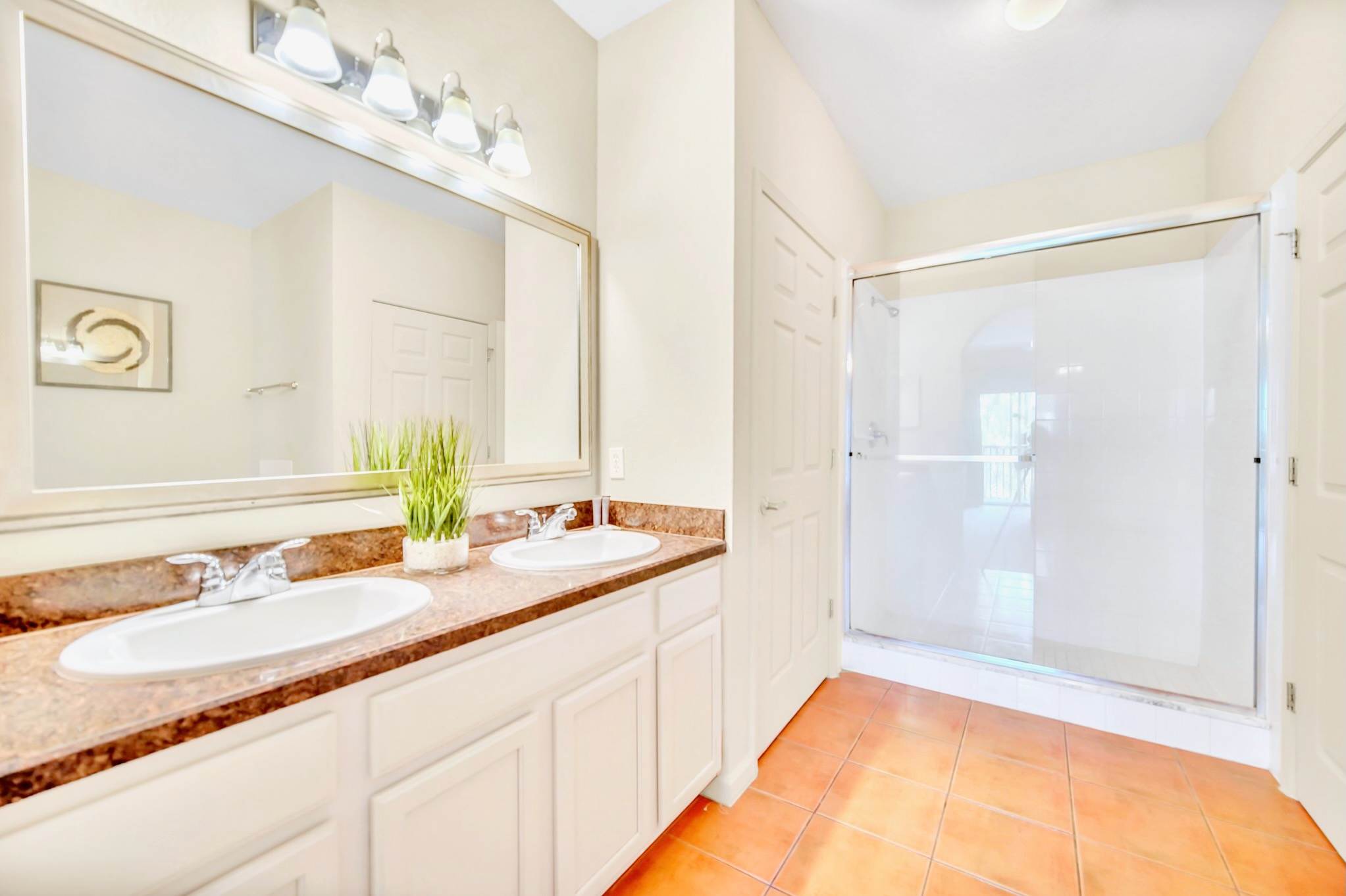 A spacious bathroom with a double vanity featuring white sinks and dark granite countertops, a large mirror with vanity lighting, and a glass-enclosed walk-in shower. Terracotta-colored tile flooring is visible.