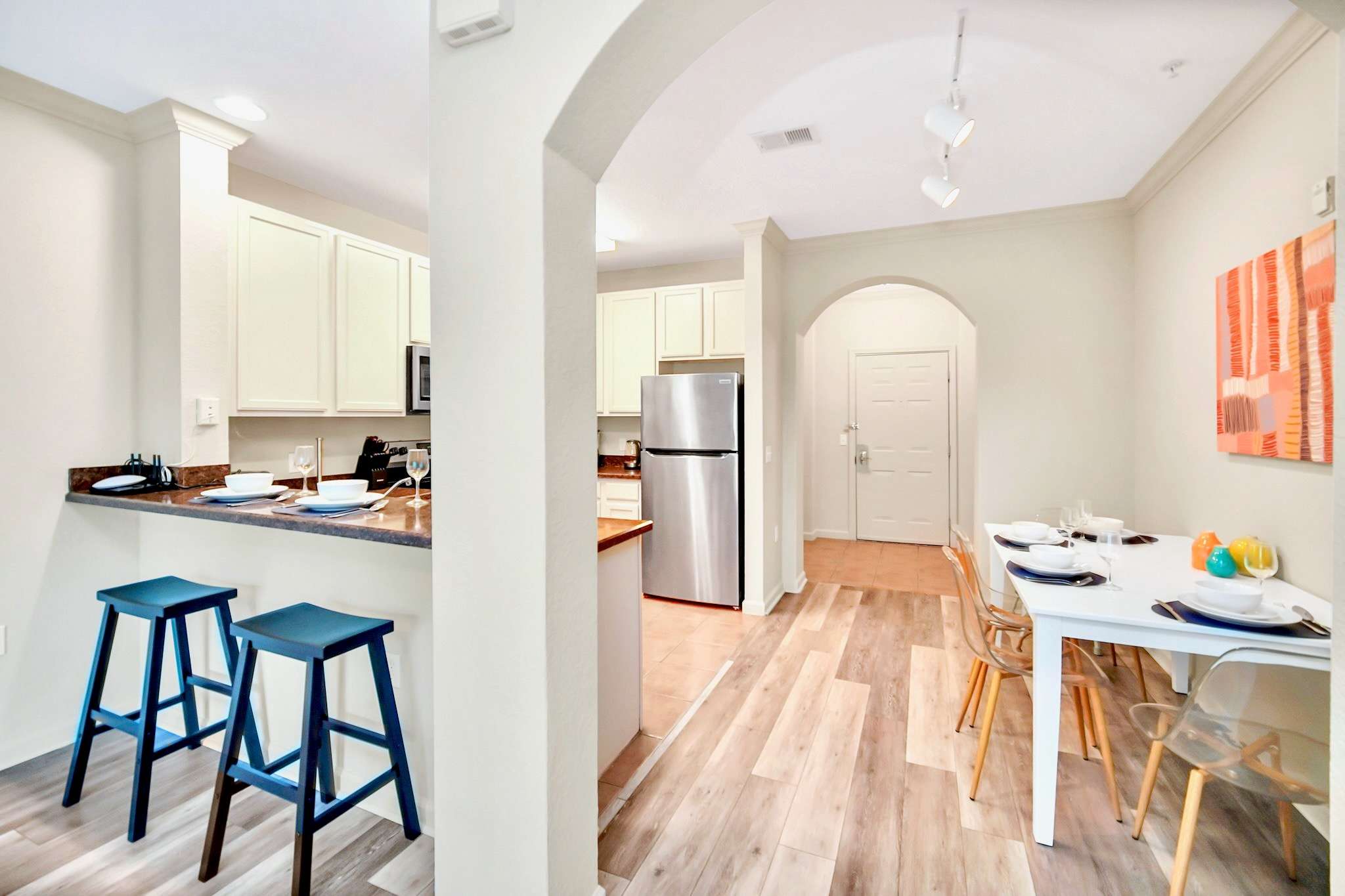 A modern open-concept space featuring a kitchen island with two blue bar stools, a dining area with a white table and clear chairs, and light wood-look flooring. Arched doorways lead to other areas.