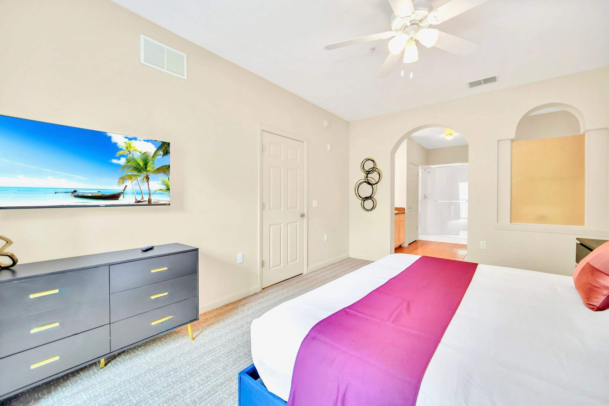 A spacious bedroom with a king-sized bed featuring a dark blue frame and purple and white bedding, a wall-mounted TV, and a dark gray dresser with gold accents. An arched doorway leads to an en-suite bathroom.