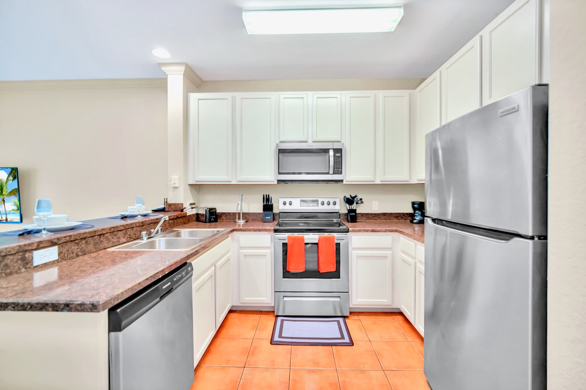 A U-shaped kitchen with white shaker cabinets, dark granite countertops, stainless steel appliances including a refrigerator, microwave, and electric range, and terracotta-colored tile flooring.