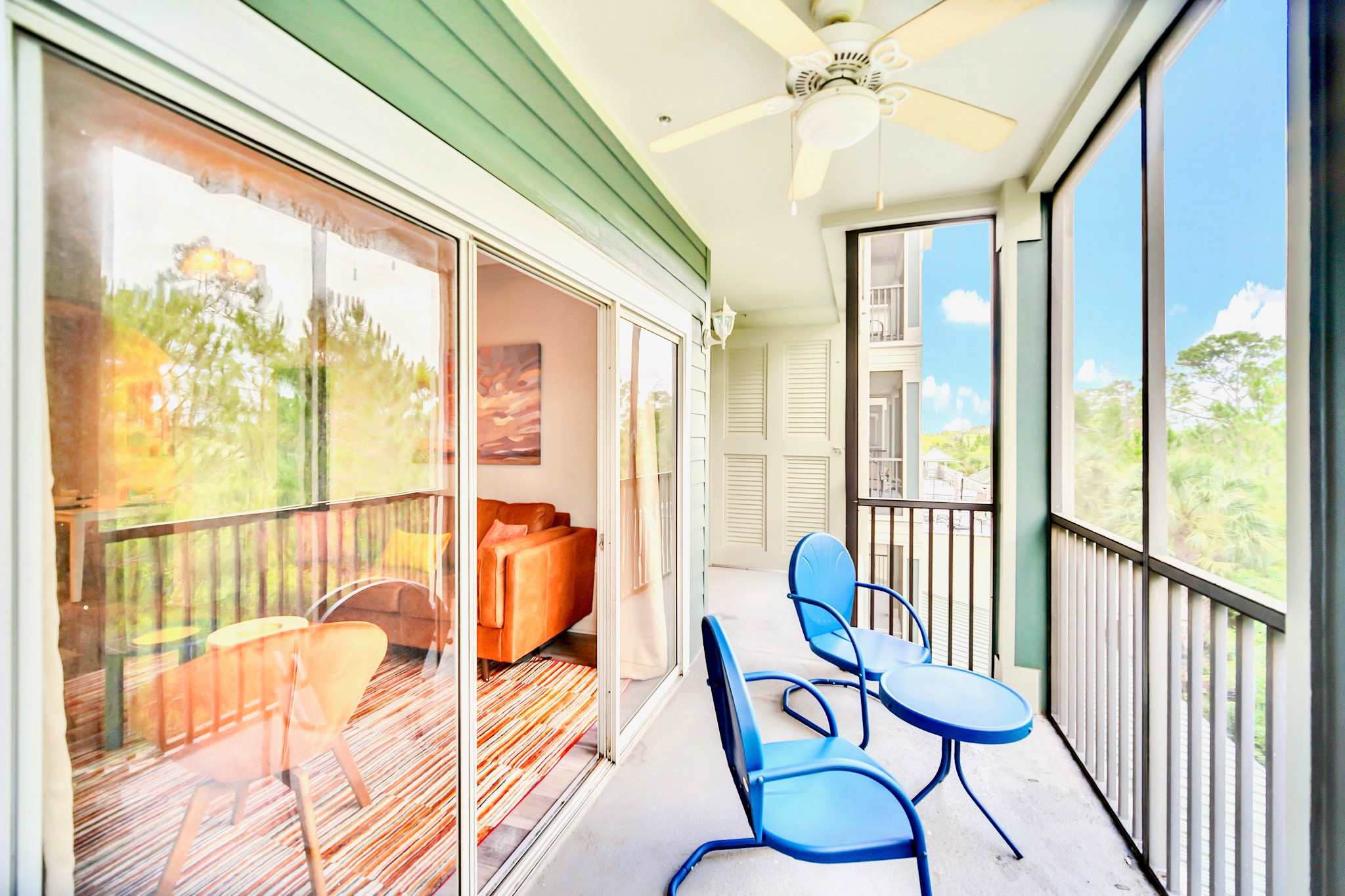 A screened-in balcony with light blue metal chairs and a small table, offering views of lush green trees and other buildings, accessible via a sliding glass door from the interior.