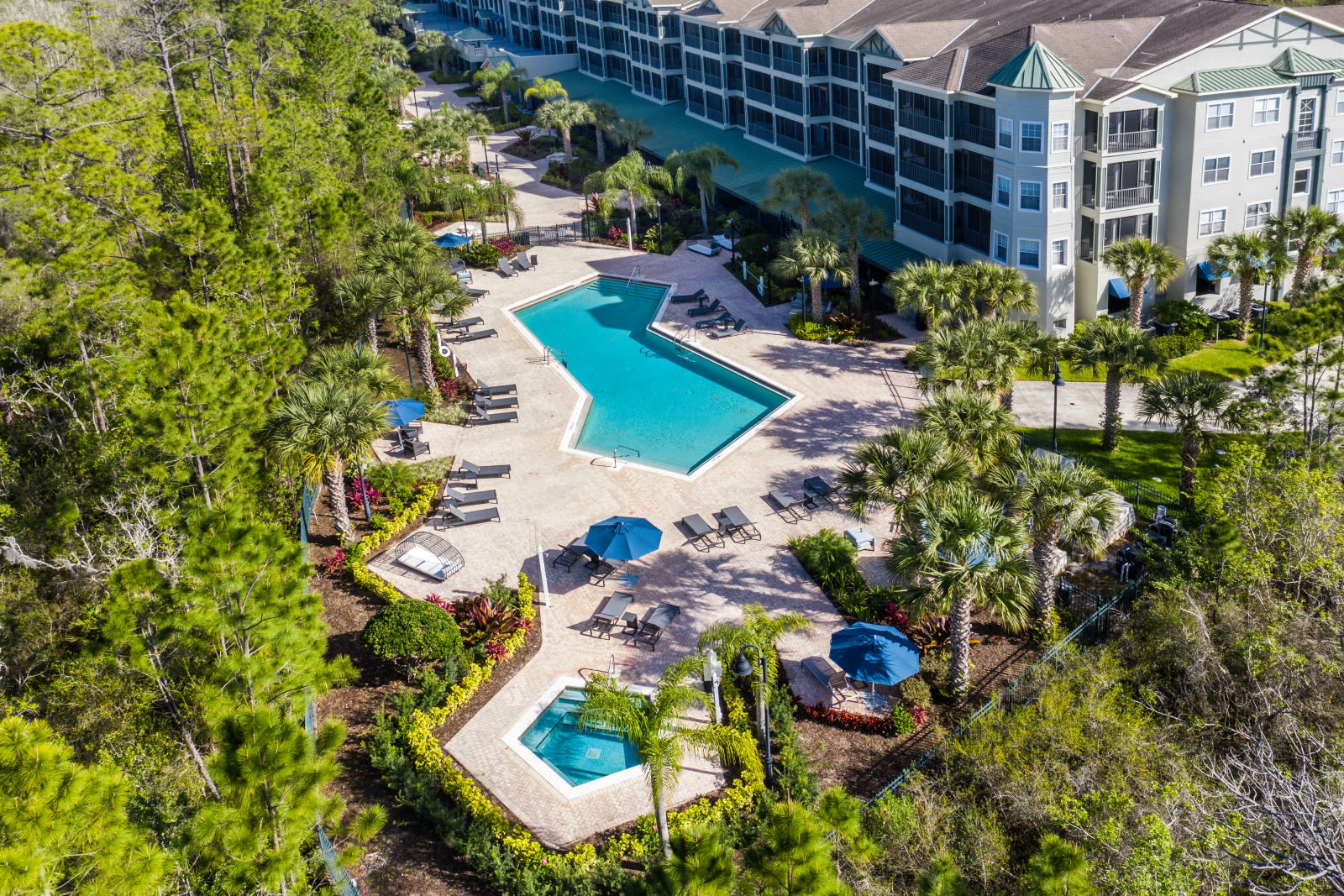 An aerial view showcasing the extensive condominium complex, featuring multiple buildings, a large outdoor swimming pool with a hot tub, and numerous lounge chairs, all nestled within a lush, treed landscape.
