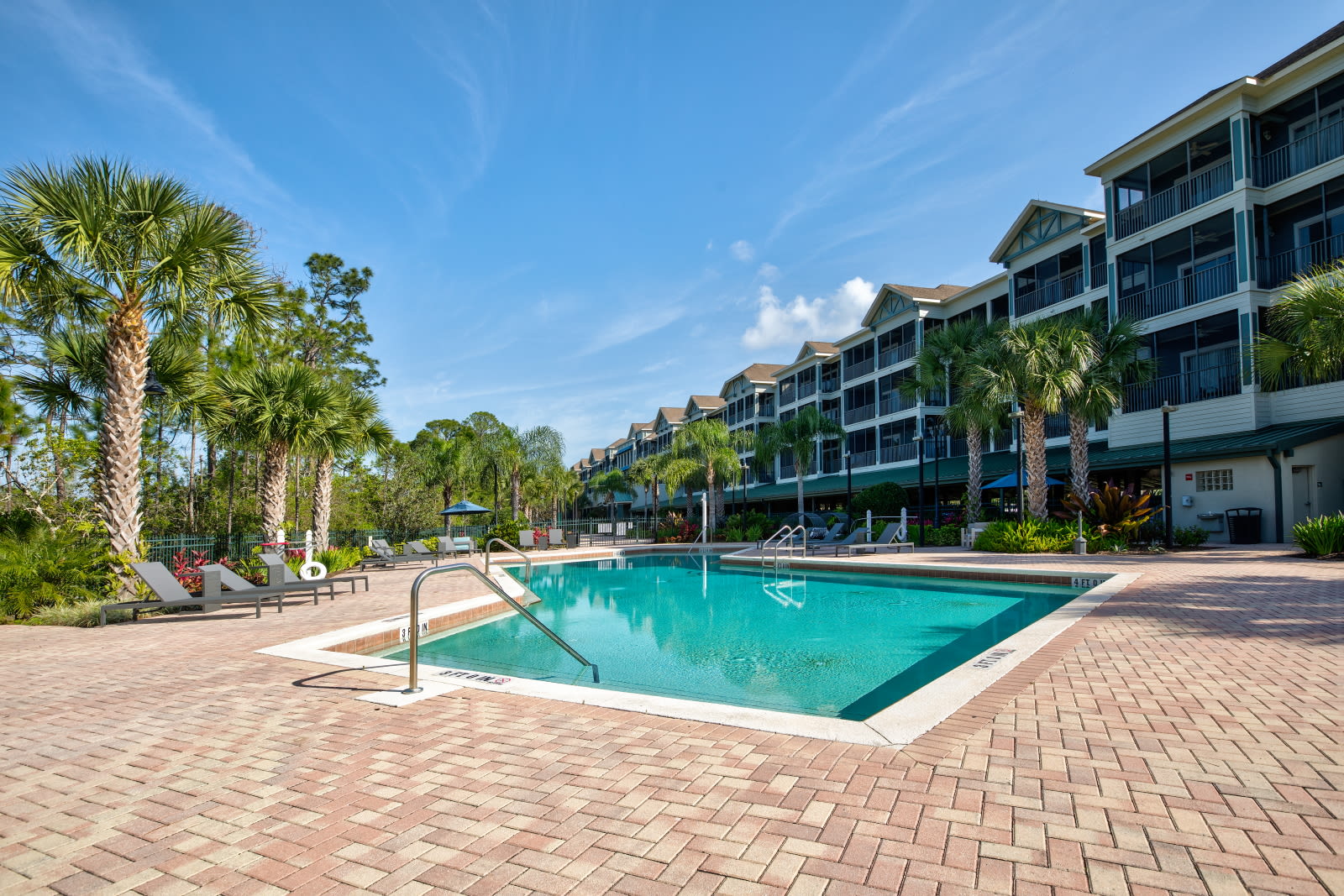 A spacious outdoor swimming pool with clear blue water, surrounded by brick paving and numerous lounge chairs, set against the backdrop of the multi-story condominium buildings and palm trees.