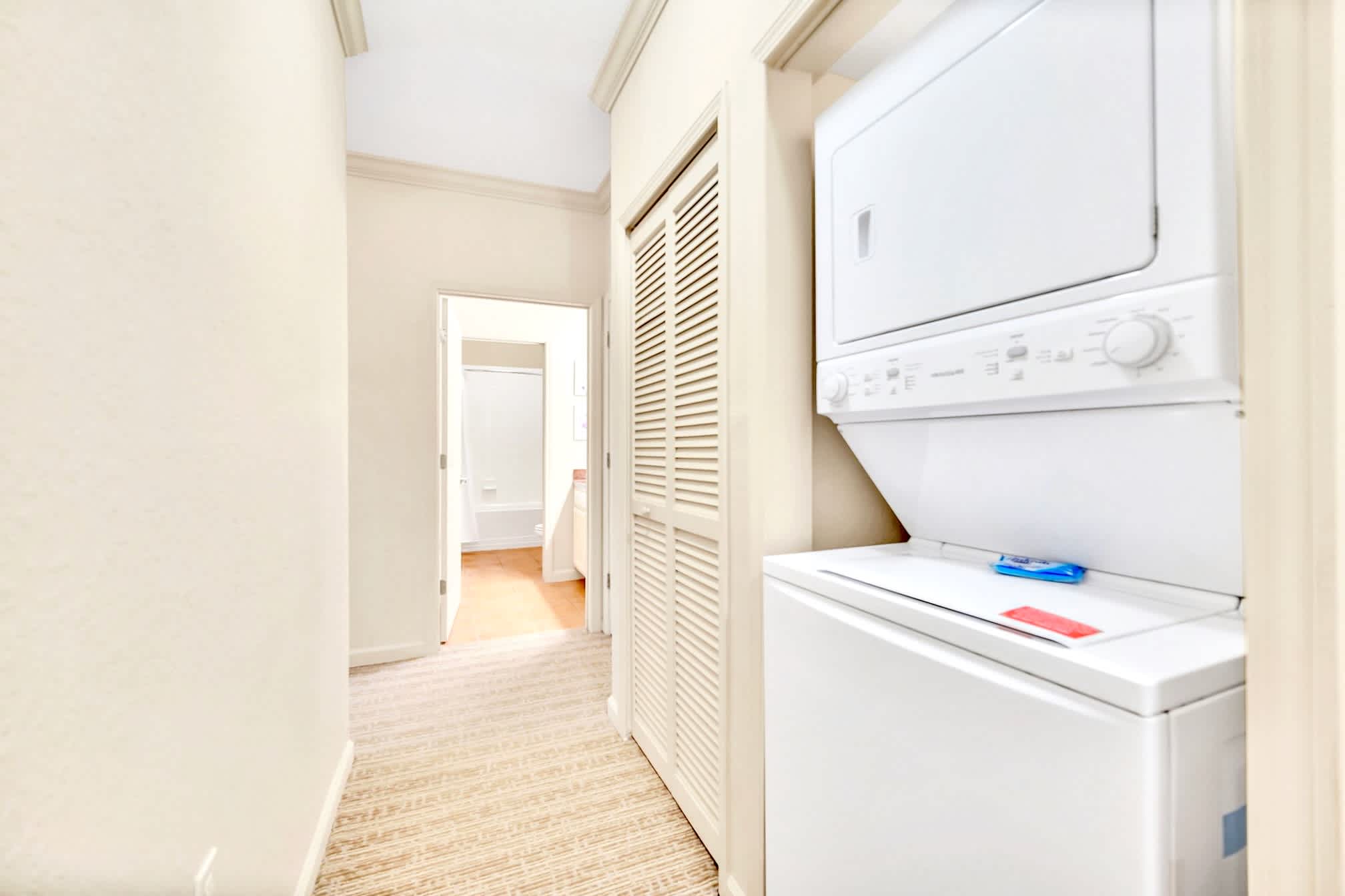 A narrow hallway featuring a stacked white washer and dryer unit neatly tucked into a louvered closet, with a glimpse of a bathroom at the far end.