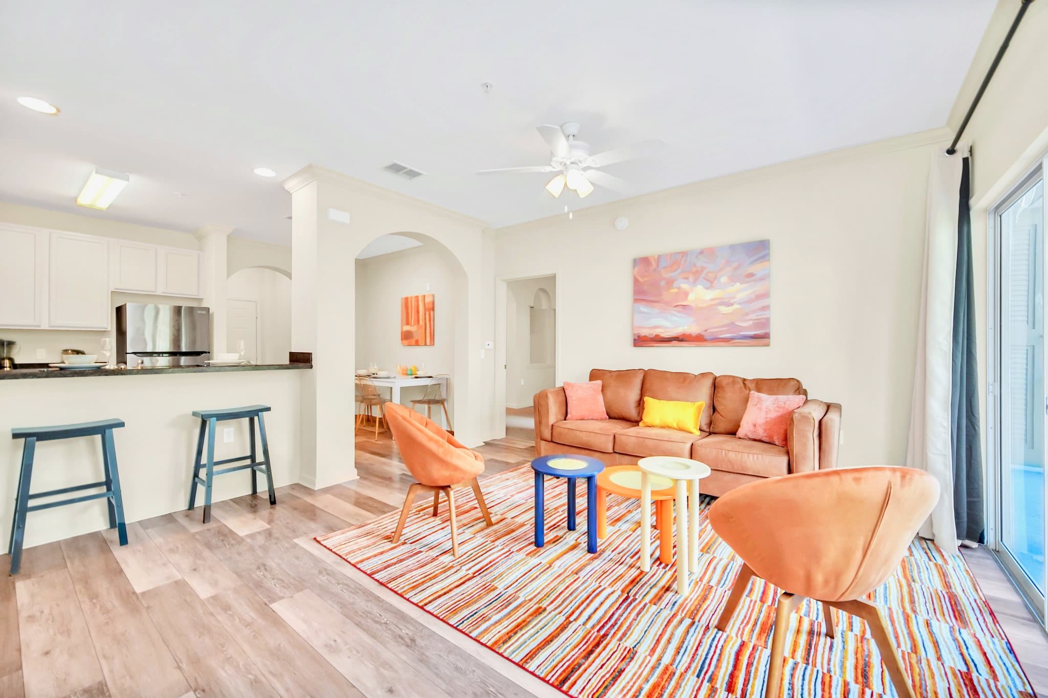 A wide shot of the open-concept living room, featuring a brown leather sofa, two orange armchairs, and colorful coffee tables on a striped rug, with the kitchen breakfast bar and dining area visible in the background.