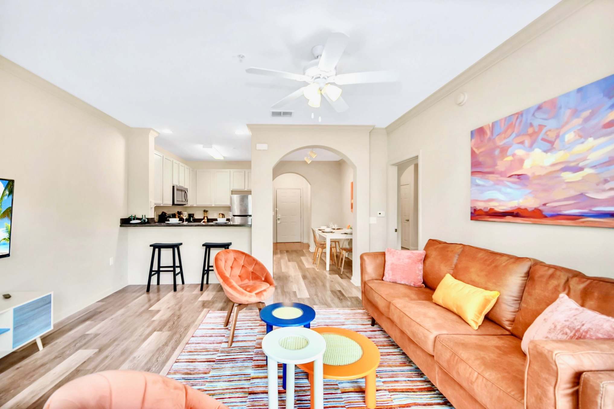 A spacious open-plan living room with a brown leather sofa, two orange velvet armchairs, and colorful round coffee tables on a striped rug, with a view towards the kitchen and dining area through an arched doorway.
