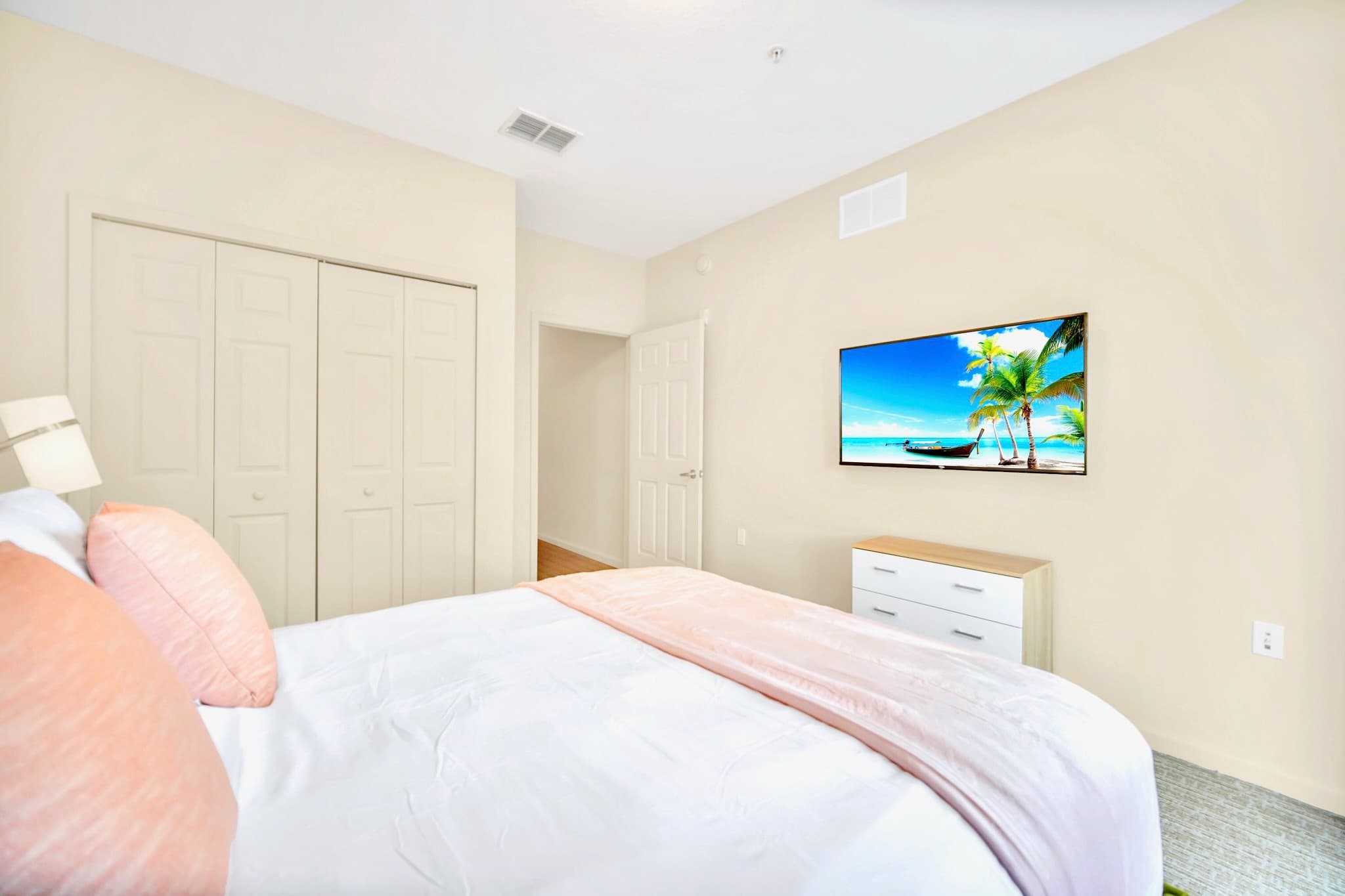 A bright bedroom with a queen-sized bed, a wall-mounted TV, and a white dresser. Double bi-fold closet doors are visible, and the room has light carpeted flooring.