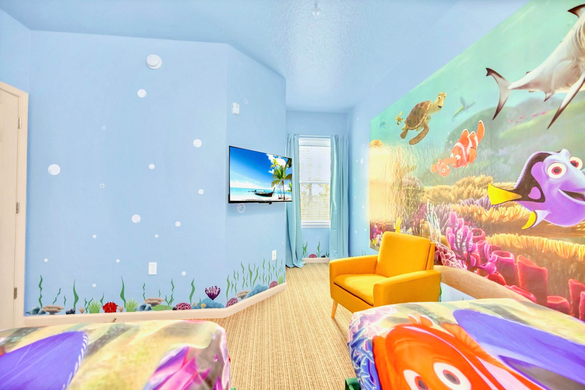 A vibrant children's bedroom featuring blue walls adorned with Finding Nemo/Dory murals and underwater decals, two beds with themed bedding, a bright yellow armchair, and a wall-mounted flat-screen TV.