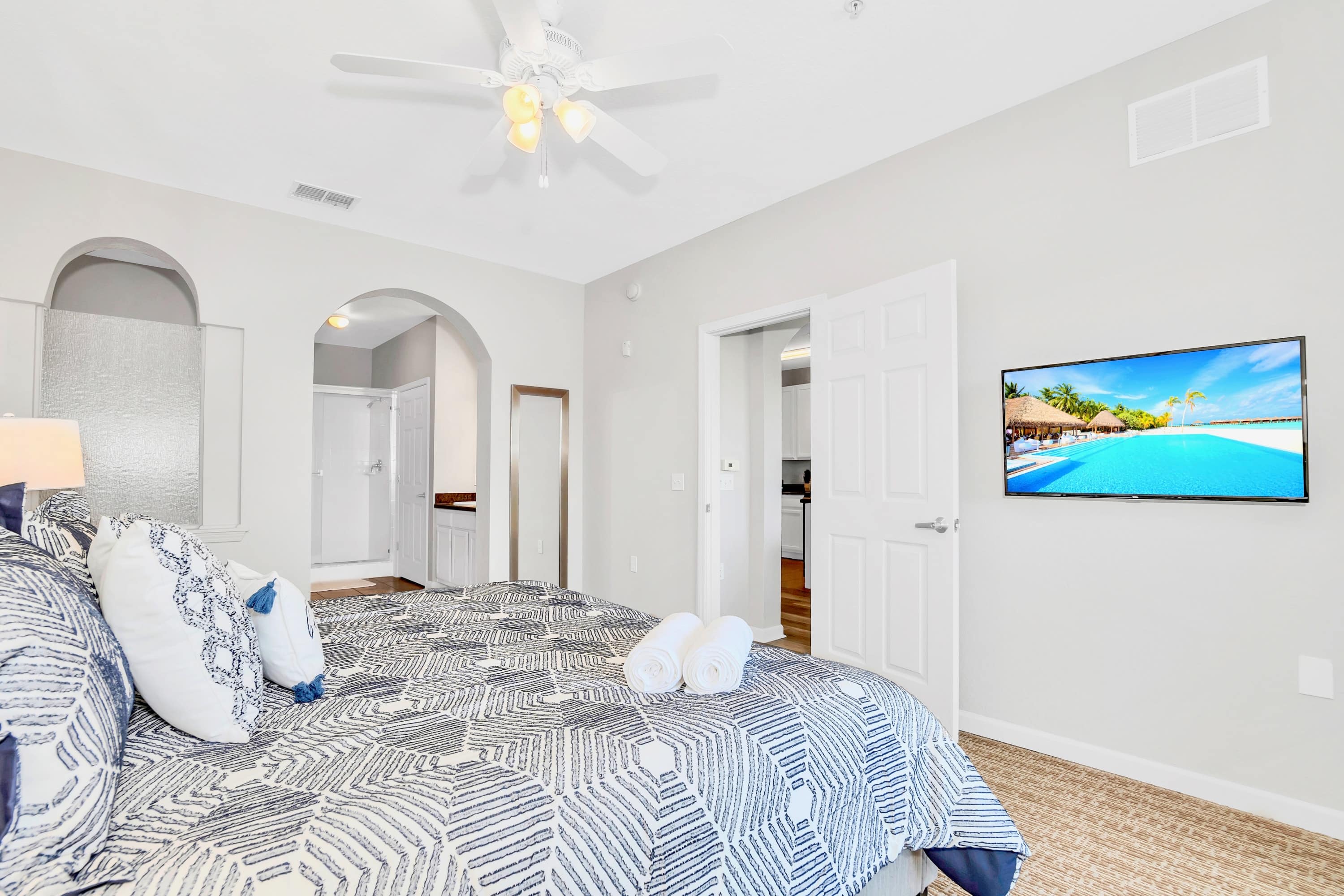 Master bedroom with a large bed, a wall-mounted TV, and an arched entryway leading to an en-suite bathroom, featuring carpeted flooring and a ceiling fan.