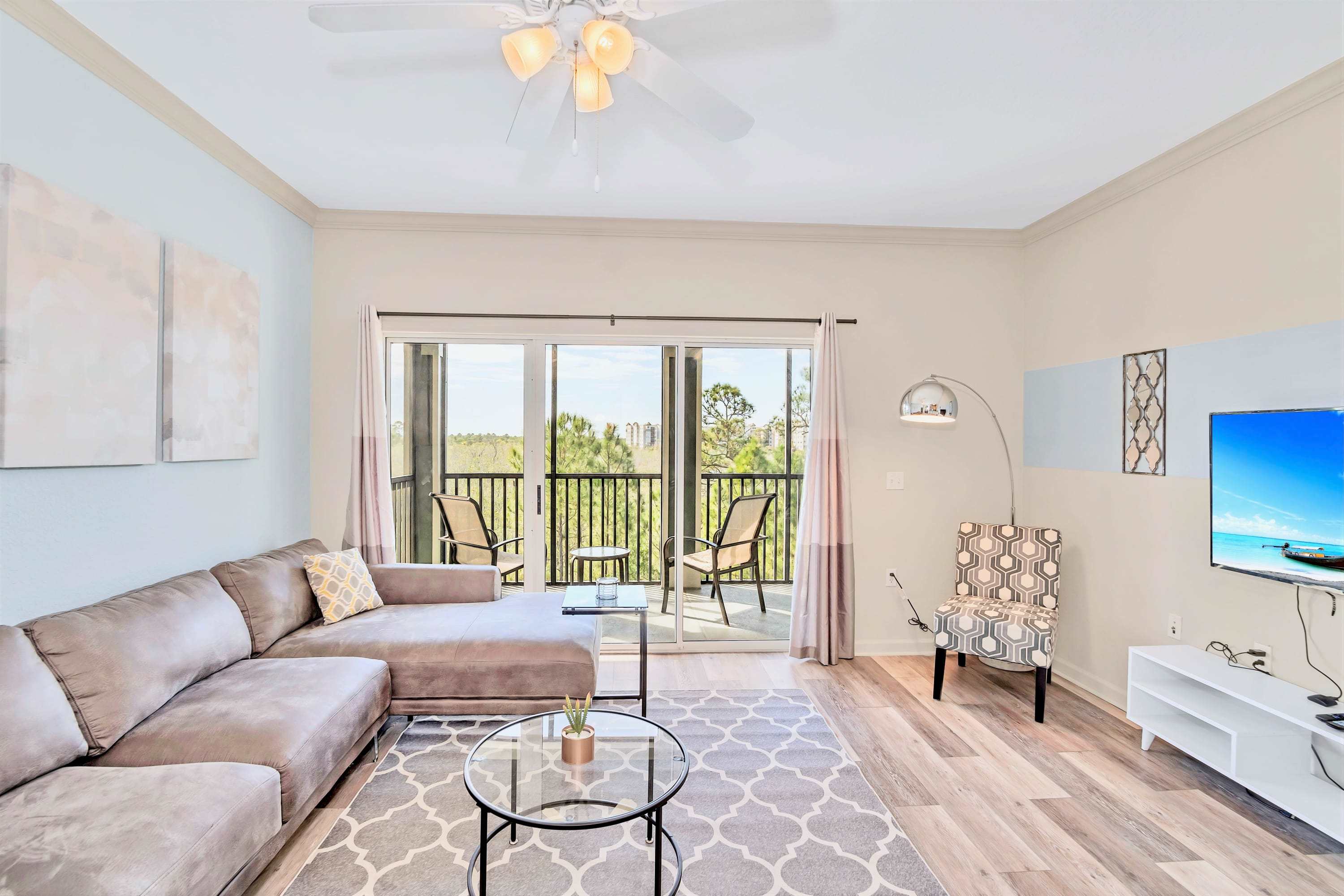 Modern living room with a large grey sectional sofa, a round glass coffee table on a geometric rug, and a wall-mounted TV, with sliding doors leading to a balcony.