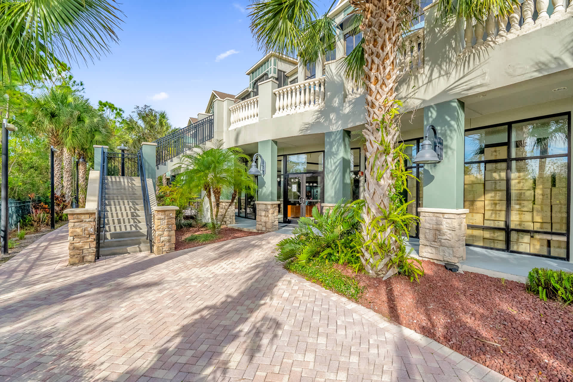 The modern entrance to a resort building, featuring a paved driveway leading to glass doors and a grand staircase with metal railings. The building has light green and grey facades, adorned with palm trees and lush landscaping.