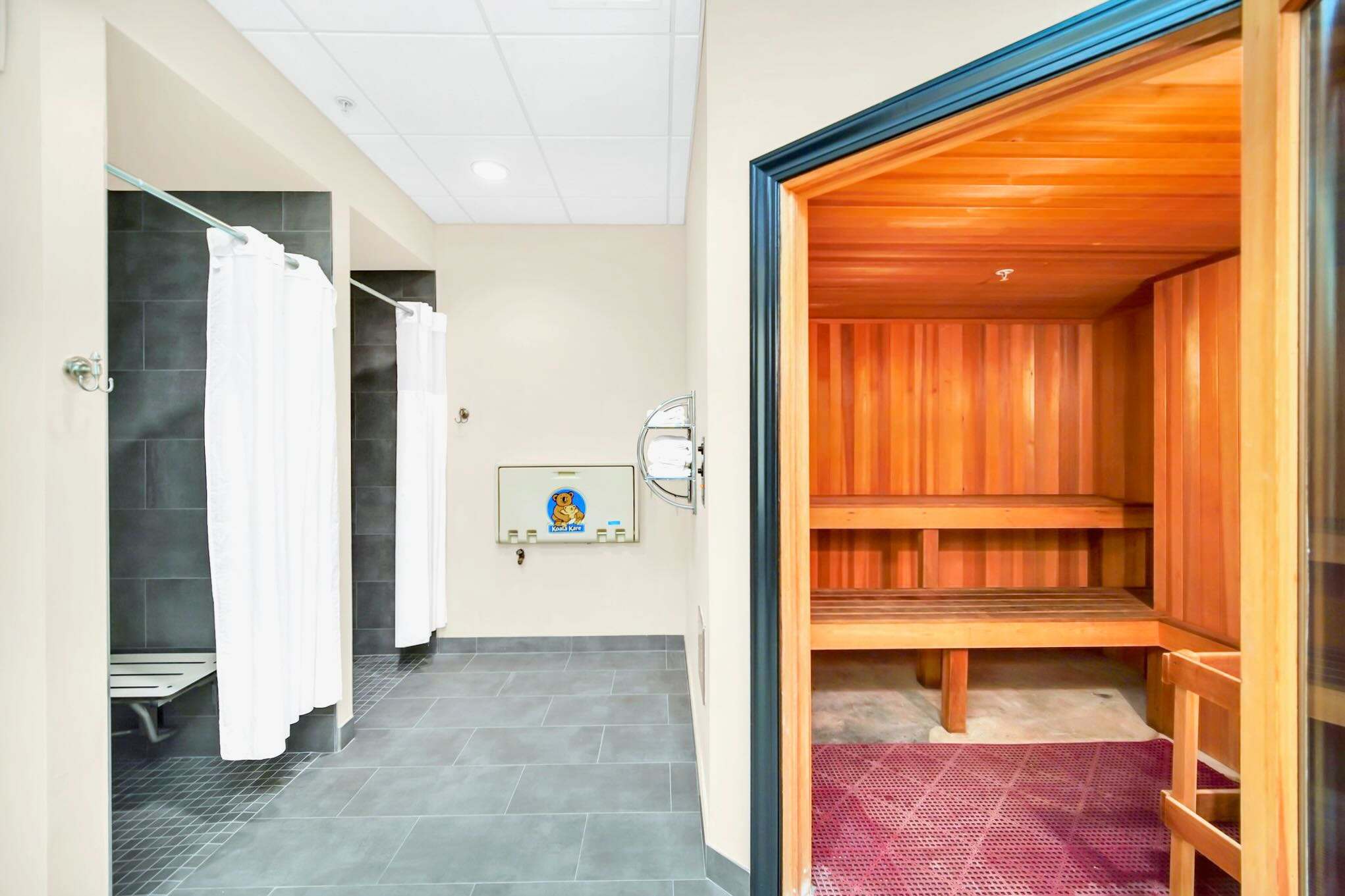 A clean, modern changing room featuring dark tiled showers with white curtains and a wooden sauna with tiered benches. A baby changing station is mounted on the wall between the shower area and the sauna entrance.
