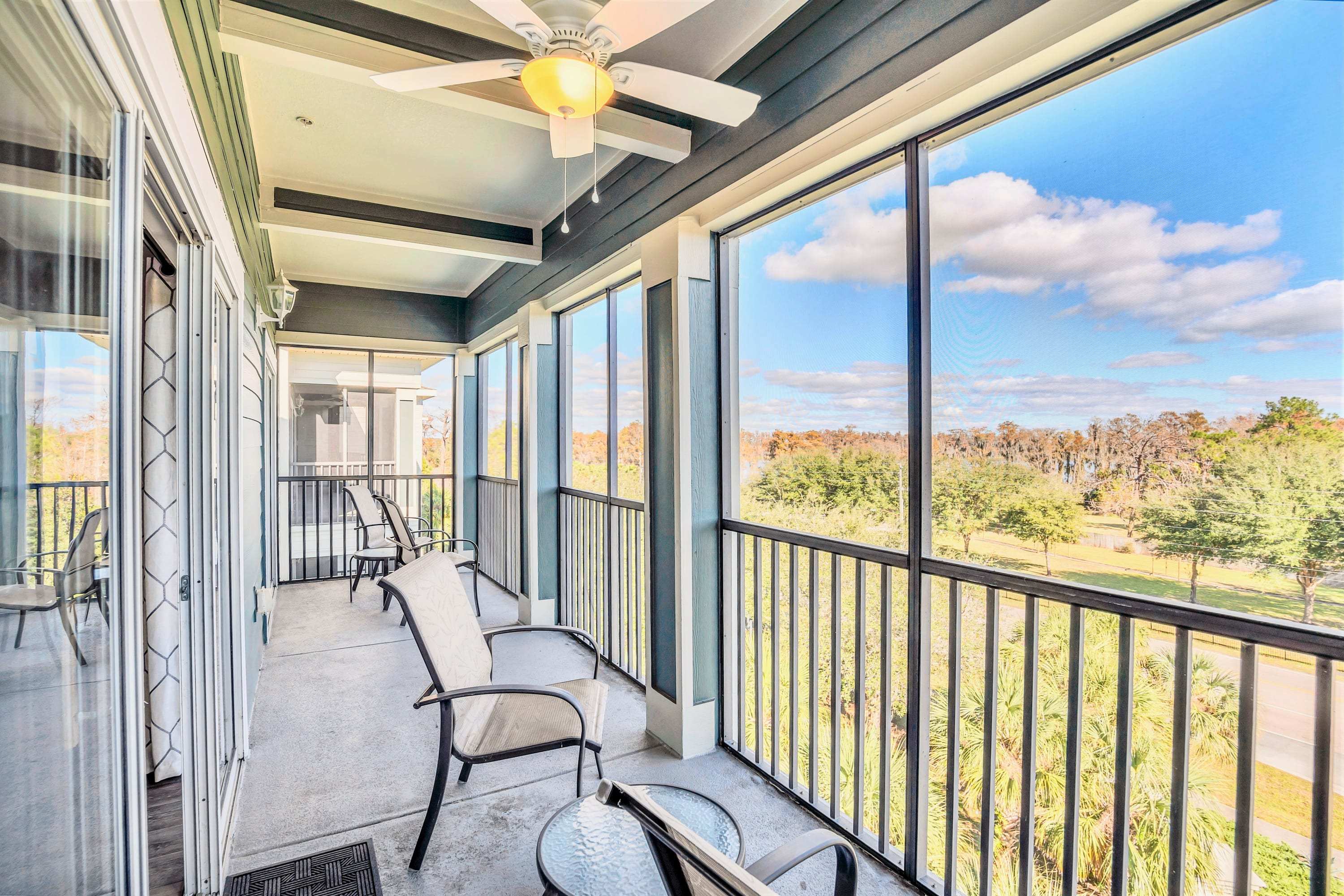 A spacious screened-in balcony with multiple outdoor chairs and small tables, offering a view of lush trees and a bright blue sky. A ceiling fan provides comfort, and the space is enclosed by dark railings and screens.
