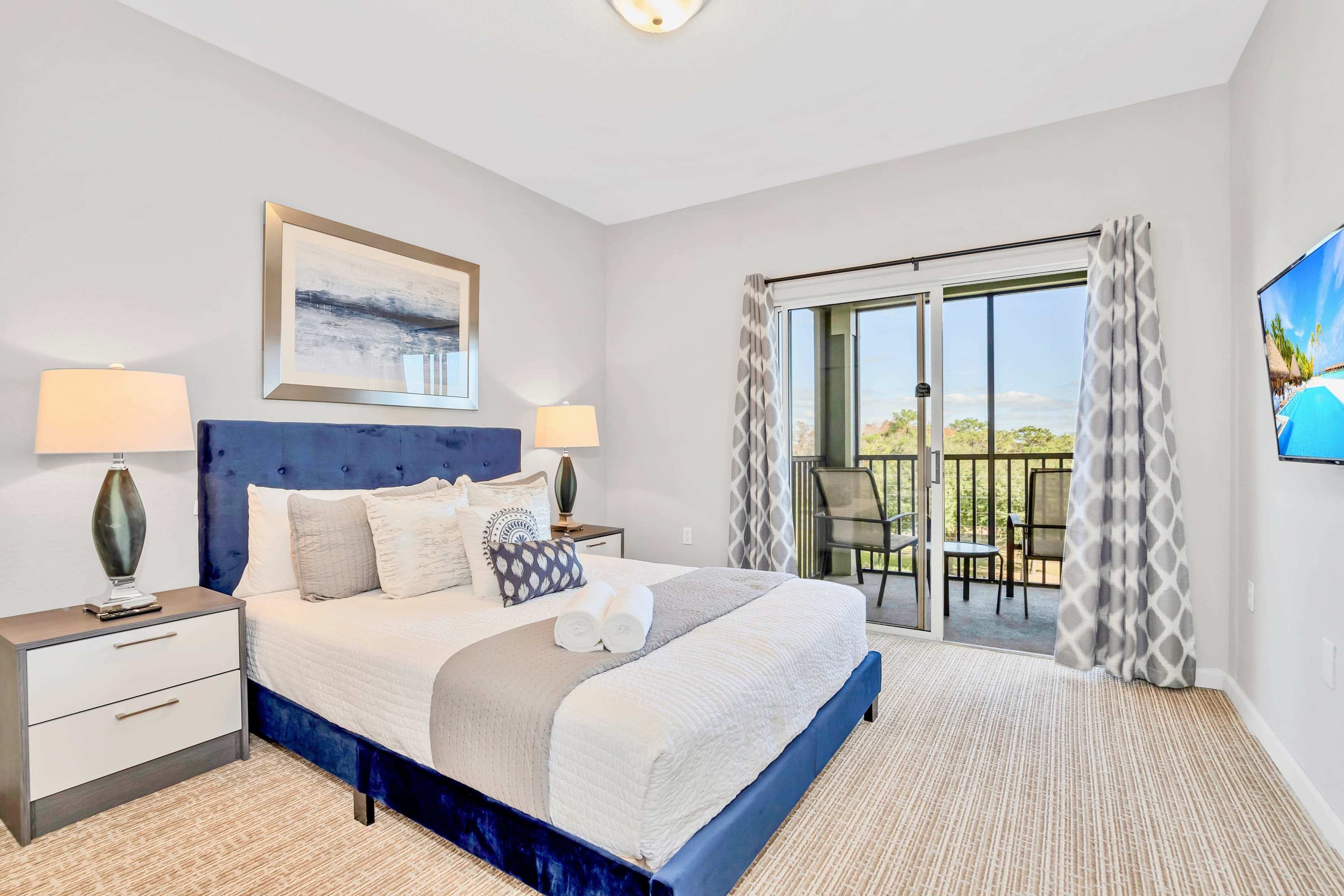 A bright bedroom with a blue velvet bed, two nightstands with lamps, and a large sliding glass door leading to a private balcony with outdoor seating. The room features light-colored carpet and a modern abstract artwork above the bed.