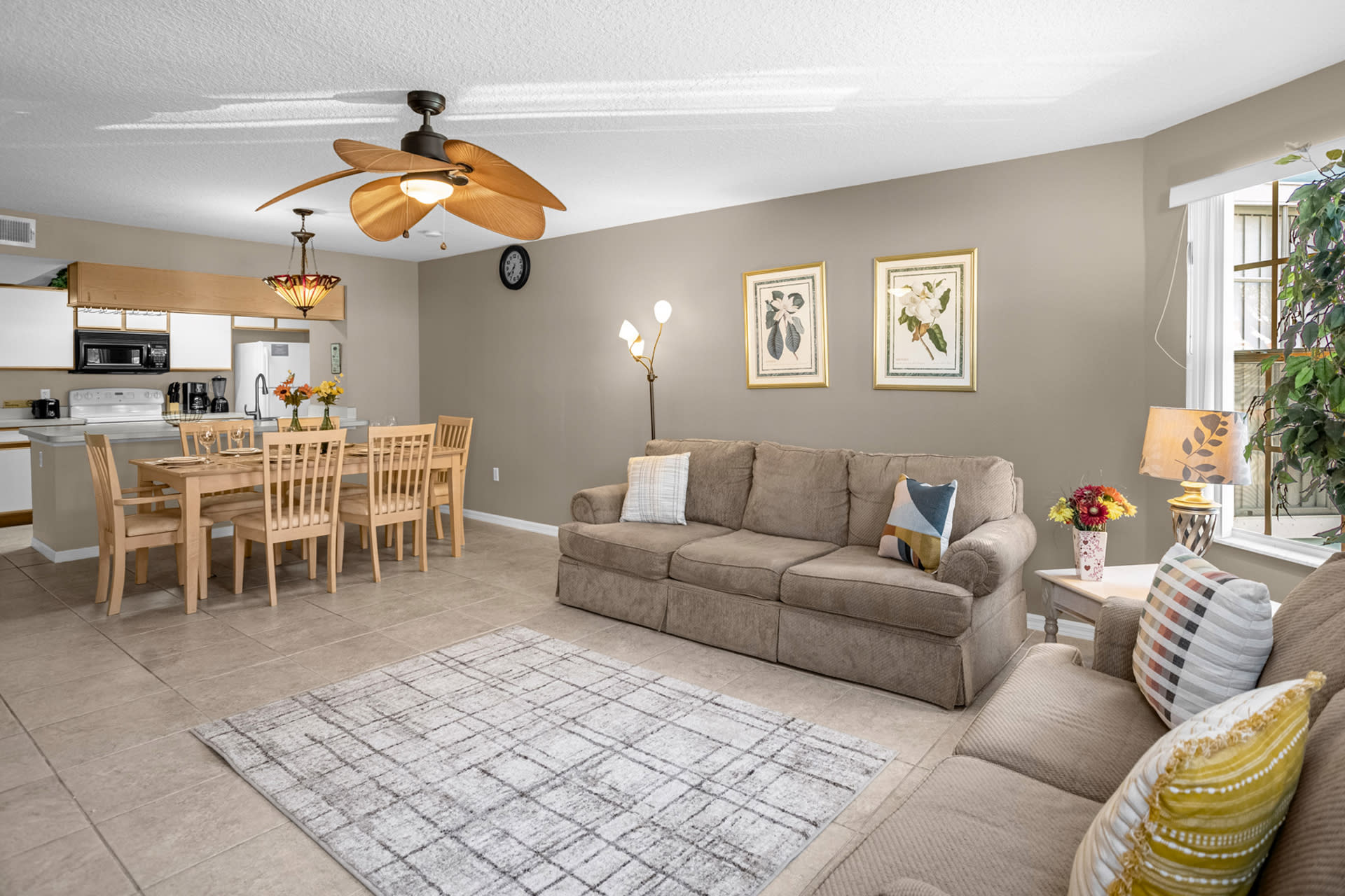 An expansive view of the open-concept living and dining area, featuring two beige sofas, a patterned rug, a dining table, and a large ceiling fan, all on light tiled flooring.