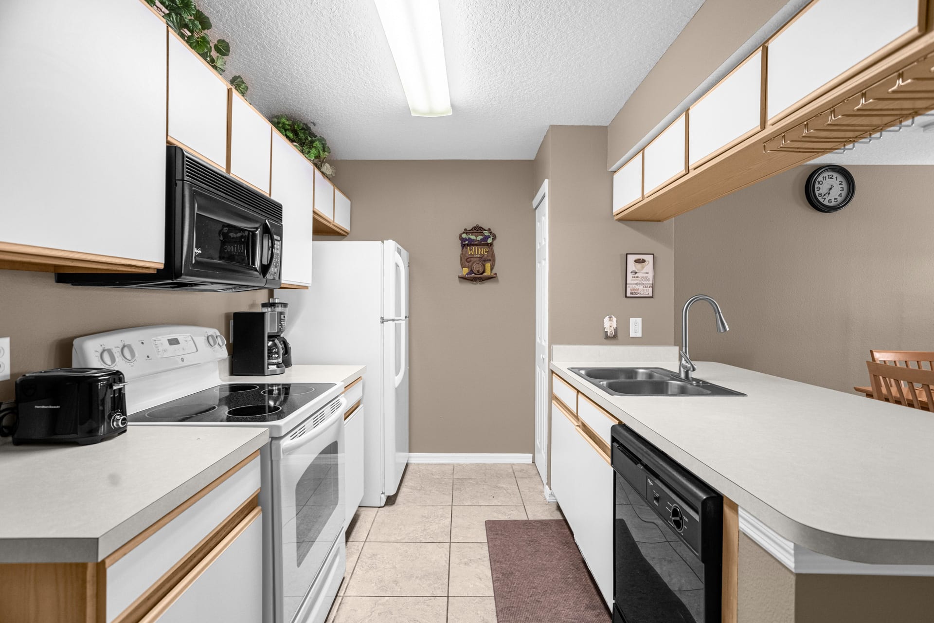 A galley-style kitchen with light beige walls, white cabinets, and white appliances including a refrigerator, electric range, and dishwasher. The space features a double sink and a wall-mounted clock.