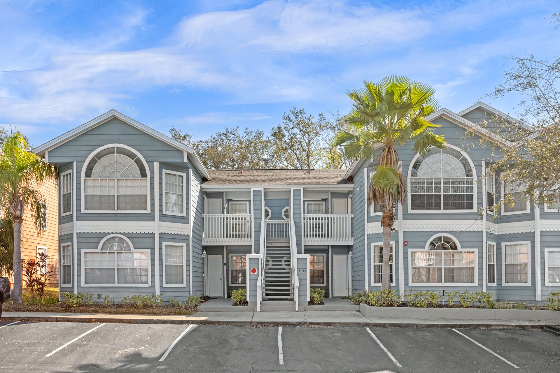 The front exterior of a light blue two-story condominium building with white trim, multiple windows, balconies, and an outdoor staircase, surrounded by parking spaces and palm trees.