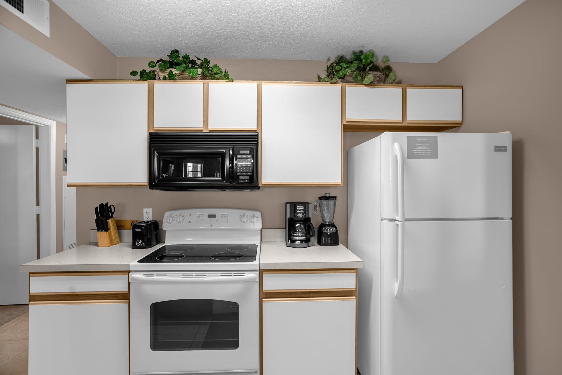 A well-lit kitchen with white upper and lower cabinets, a white electric range, and a black over-the-range microwave. A white refrigerator stands next to the counter, and decorative greenery adorns the cabinet tops.