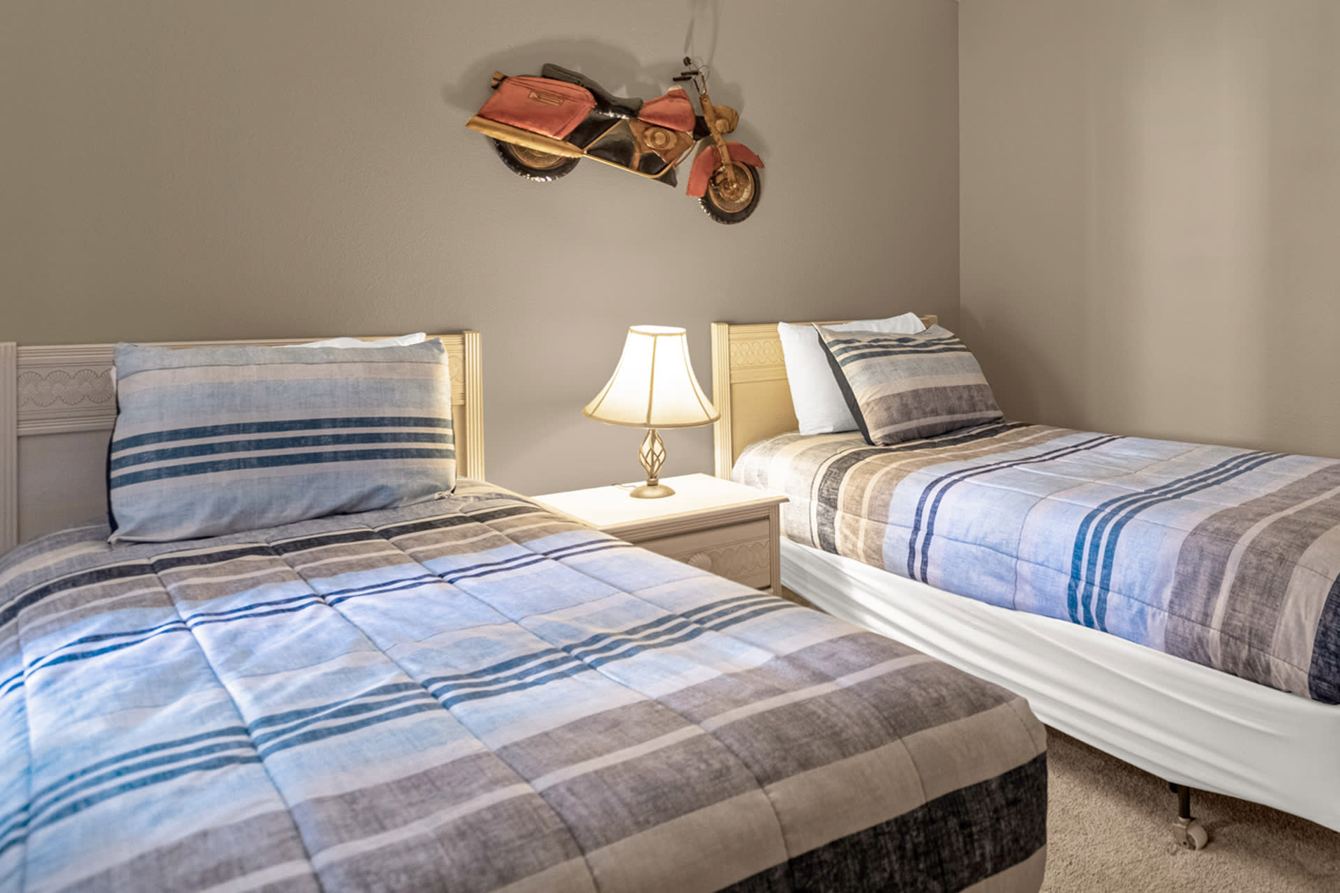 A bright bedroom featuring two twin beds with striped blue, grey, and white comforters and pillows, separated by a white nightstand with a classic lamp. A unique motorcycle wall art piece hangs above the beds on a light beige wall.