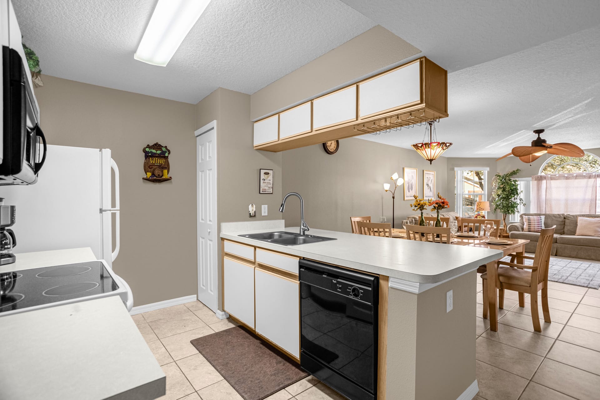 A bright kitchen with light beige walls, white cabinets, and a white countertop. It features a double sink, black dishwasher, and an open view to the dining and living areas.