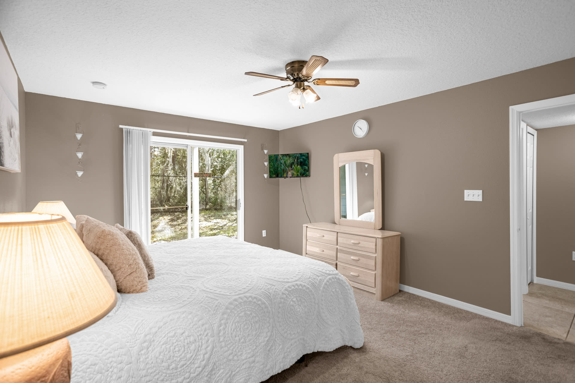 This bedroom features a queen bed with a white comforter, light brown walls, and carpeted flooring. A dresser with a mirror and a wall-mounted TV are visible, along with sliding glass doors leading to an outdoor area.