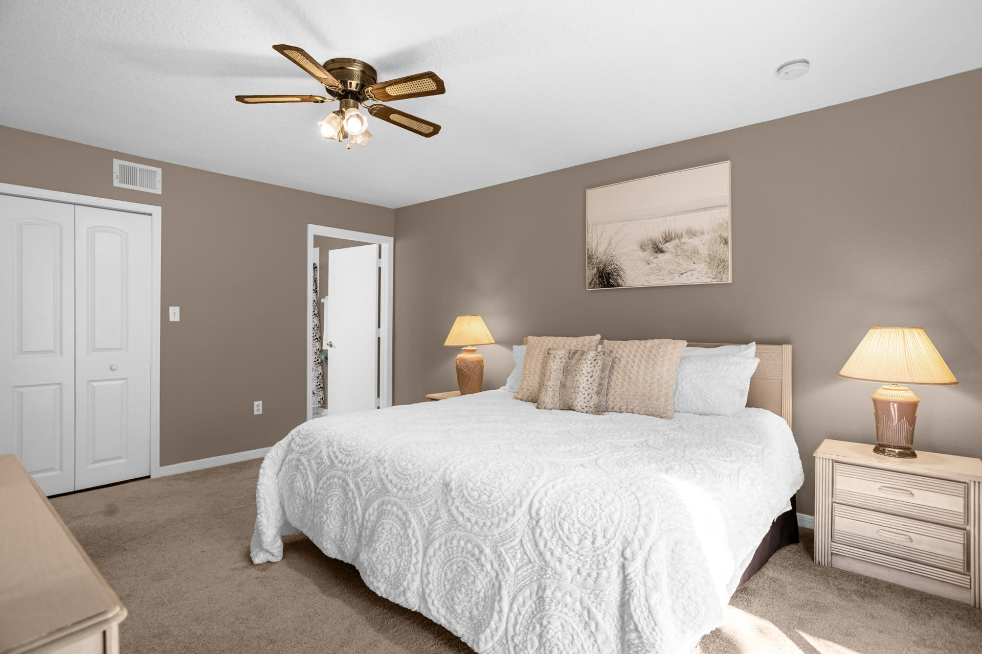 A cozy bedroom with light brown walls and carpeted flooring, featuring a queen-sized bed with a white textured comforter and decorative pillows. Two nightstands with lamps flank the bed, and a ceiling fan provides air circulation.