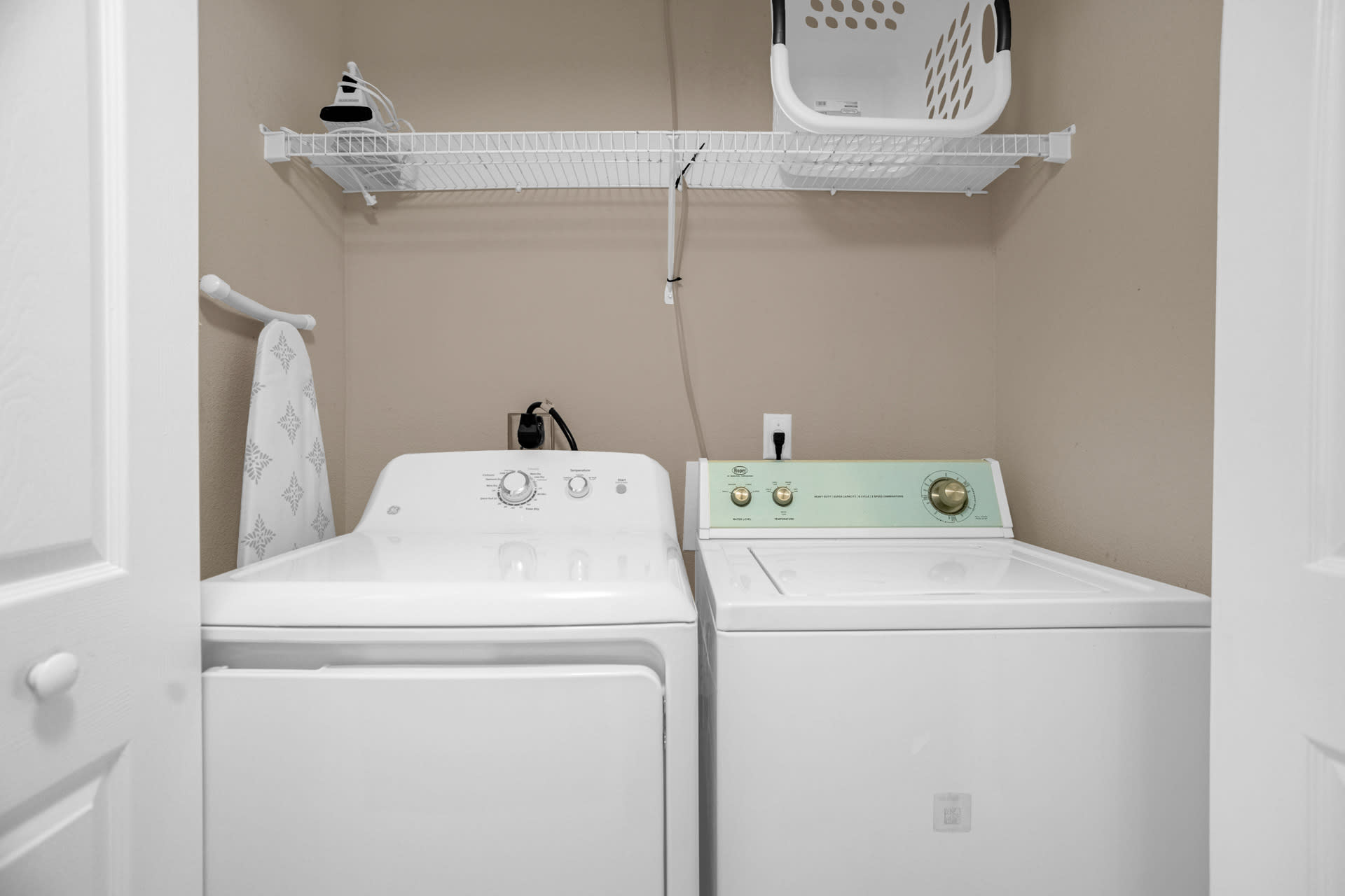 A functional laundry closet equipped with a white top-loading washing machine and a white front-loading dryer, neatly tucked under a wire shelf holding a laundry basket and an ironing board. The walls are a neutral beige.