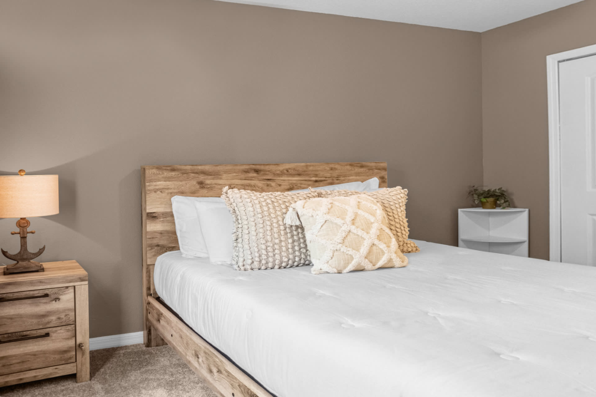 A close-up view of a queen bed with a rustic wooden frame and a white comforter, flanked by a wooden nightstand with an anchor-themed lamp. The room has light brown walls and carpeted flooring.