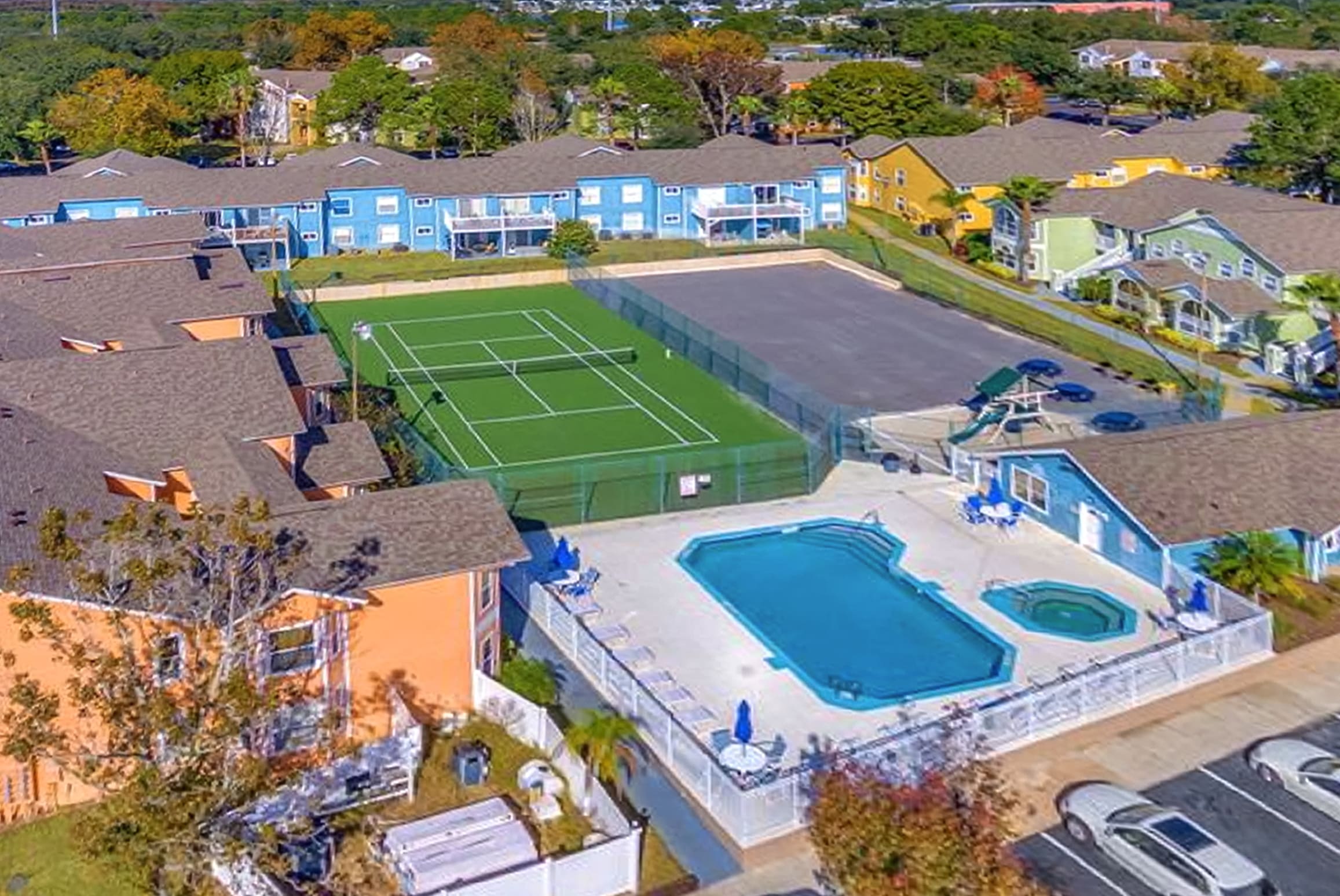 An expansive aerial view of a colorful resort community featuring a large rectangular swimming pool with a hot tub, a tennis court, and a playground, surrounded by multi-hued condominium buildings and ample parking.